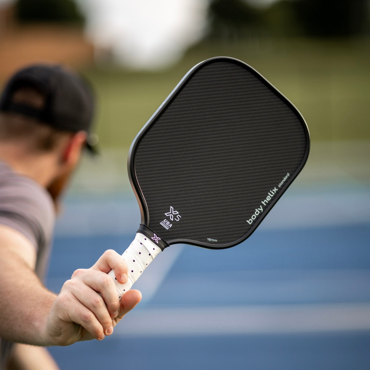 Close-up of a person holding a black Body Helix X5 Standard pickleball paddle with a white grip.