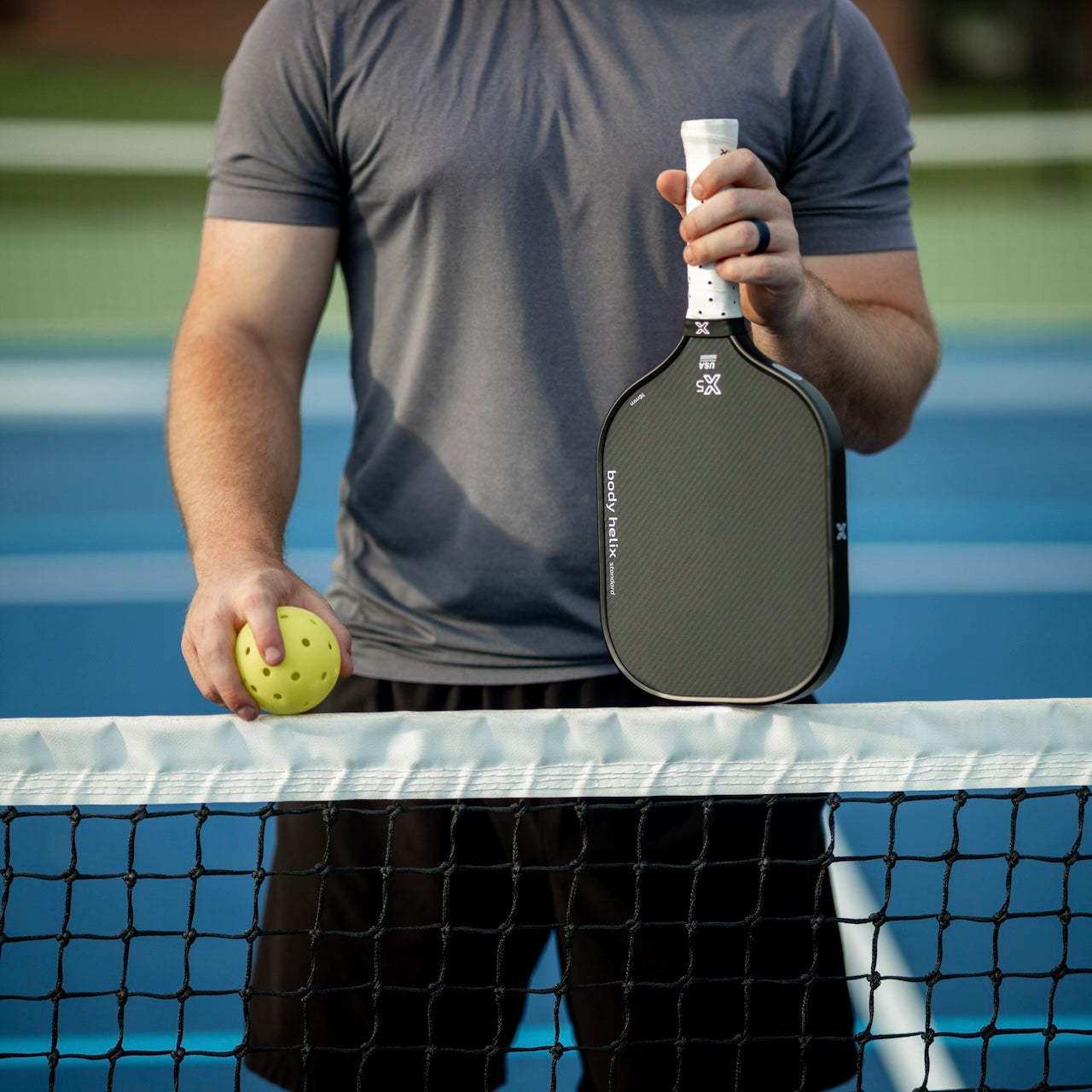 Man in gray shirt holding pickleball paddle and yellow pickleball over net.