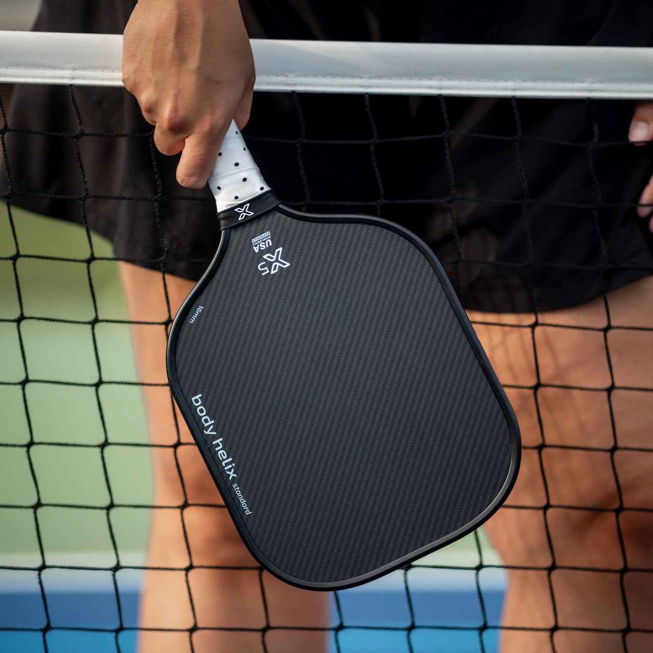 Close-up of a black pickleball paddle held in front of a net.