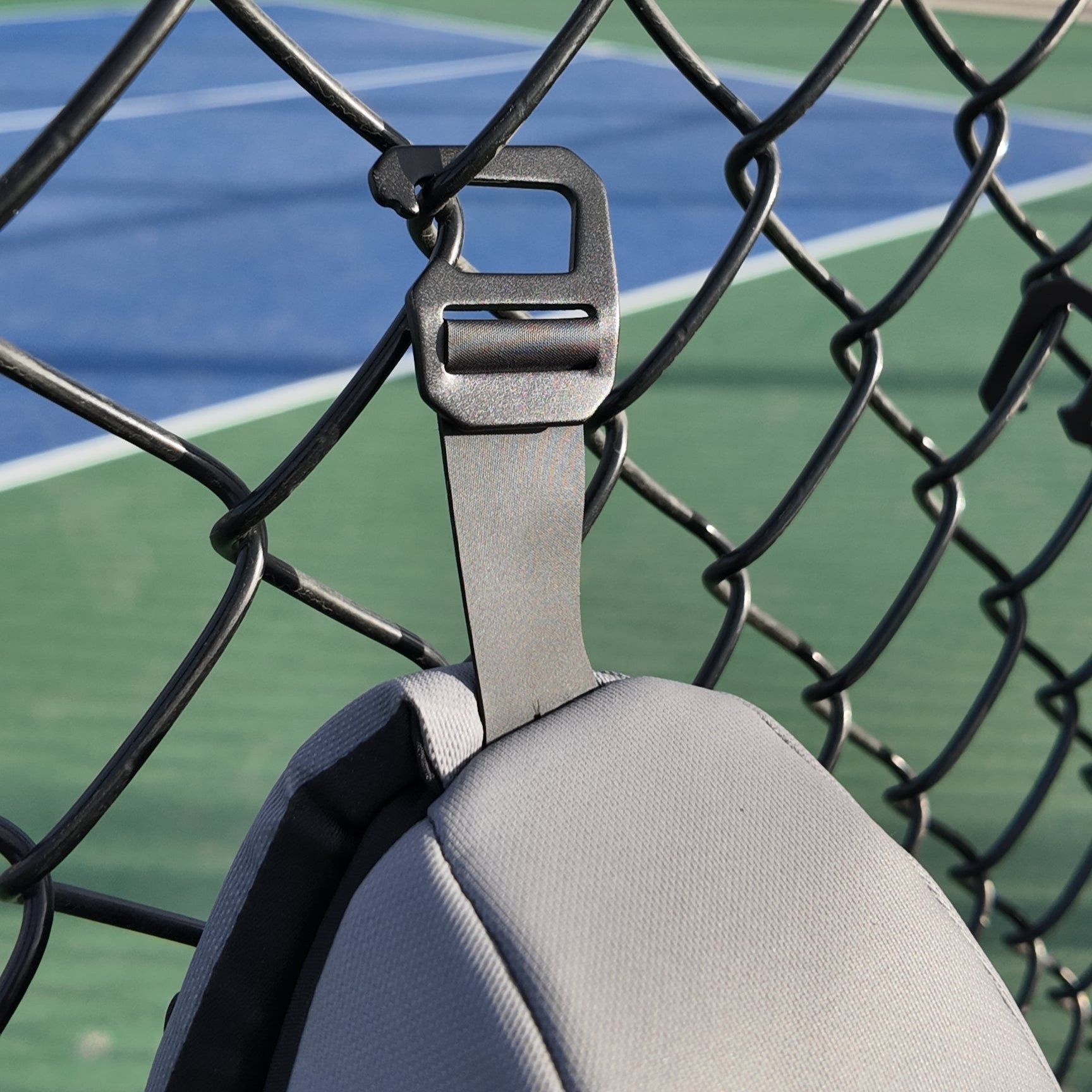 Gray pickleball sling by FORWRD, with a dark textured buckle, hanging on a black chain-link fence above a blue and green court.