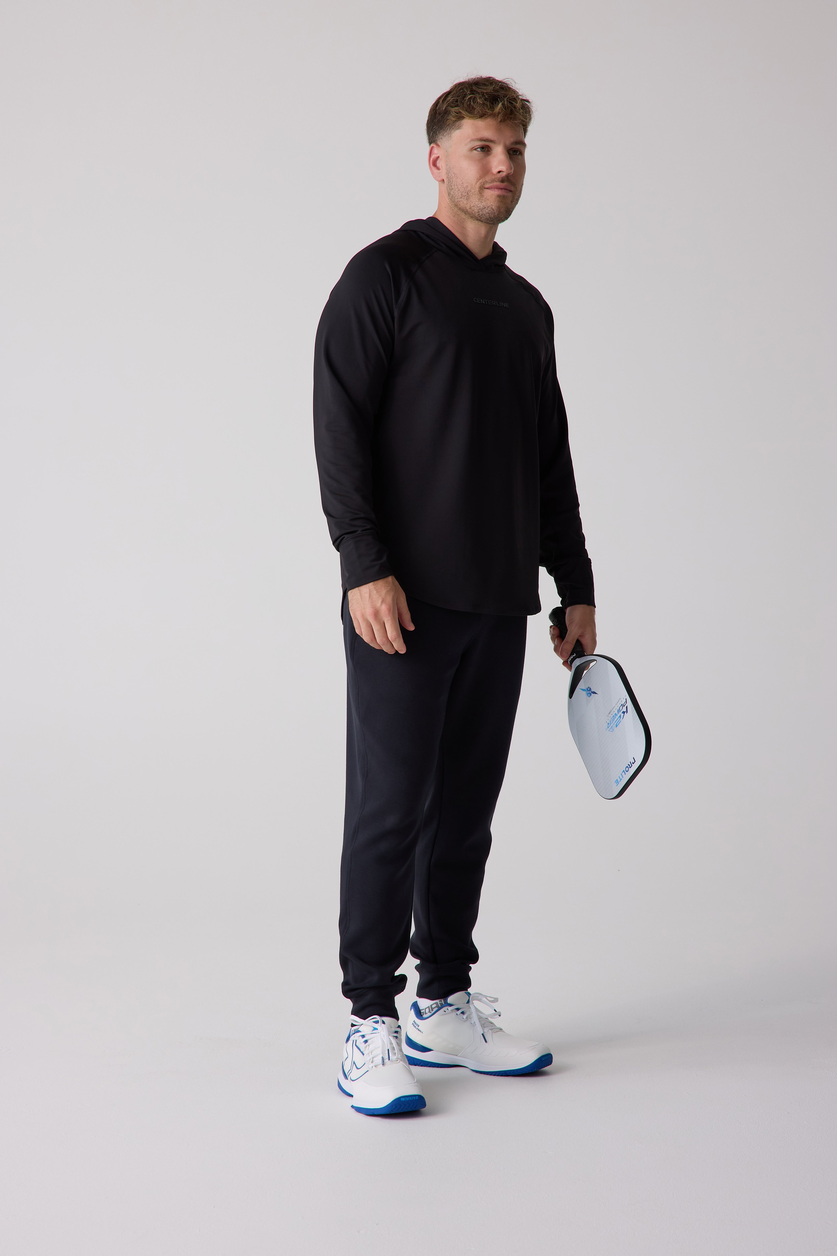 Black men's layering hoodie by Centerline Athletics with a subtle chest logo, worn by a man holding a white pickleball paddle.