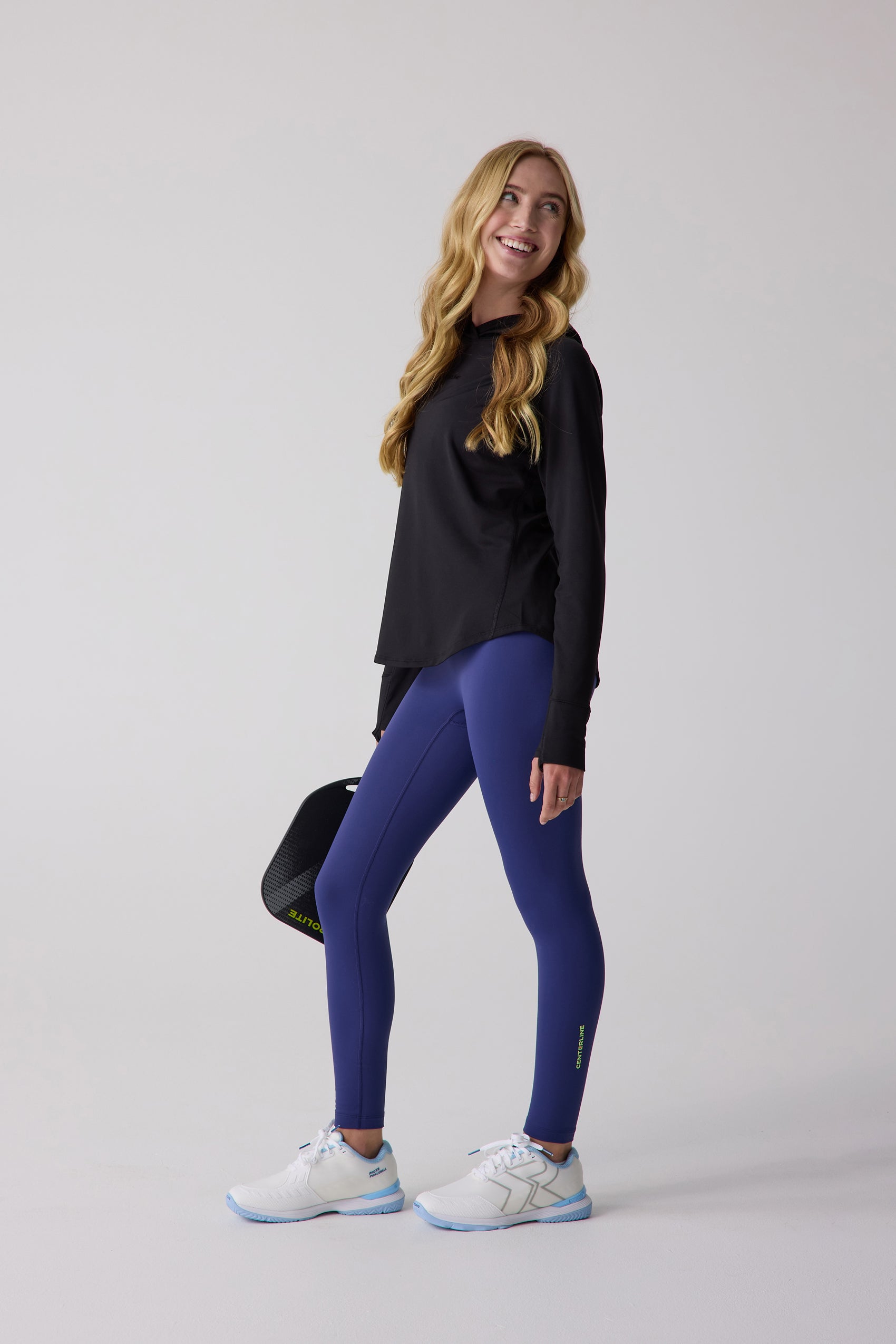 Black women's layering hoodie by Centerline Athletics, long-sleeved, worn by a smiling woman in blue leggings and white sneakers, holding a pickleball paddle.