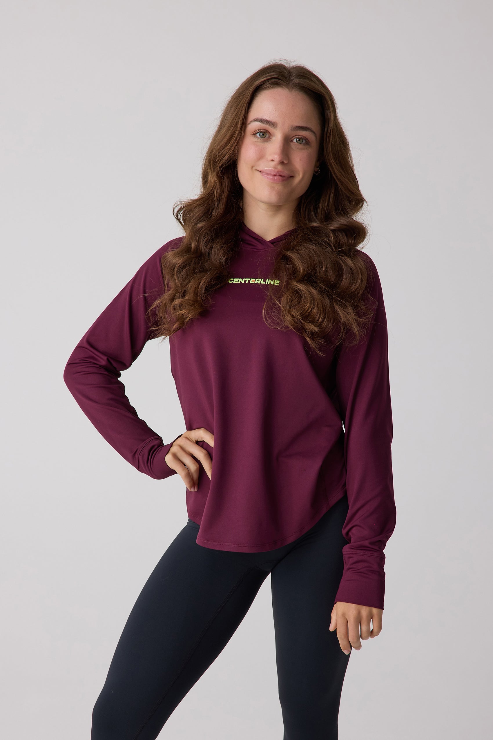 Women's burgundy long-sleeved hooded layering hoodie by Centerline Athletics, with yellow-green "CENTERLINE" text, worn with black leggings.