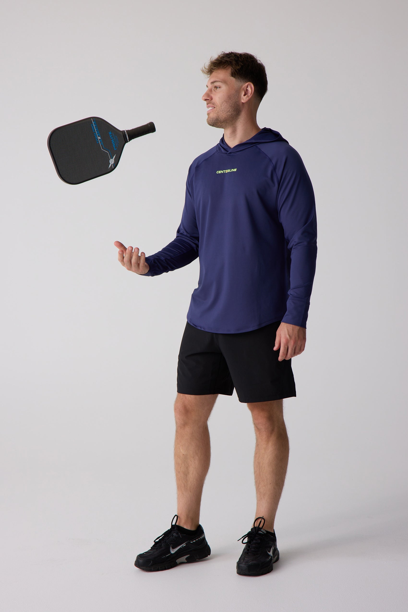 Navy blue long-sleeve men's layering hoodie by Centerline Athletics with a yellow logo, on a man wearing black shorts and sneakers, tossing a pickleball paddle.