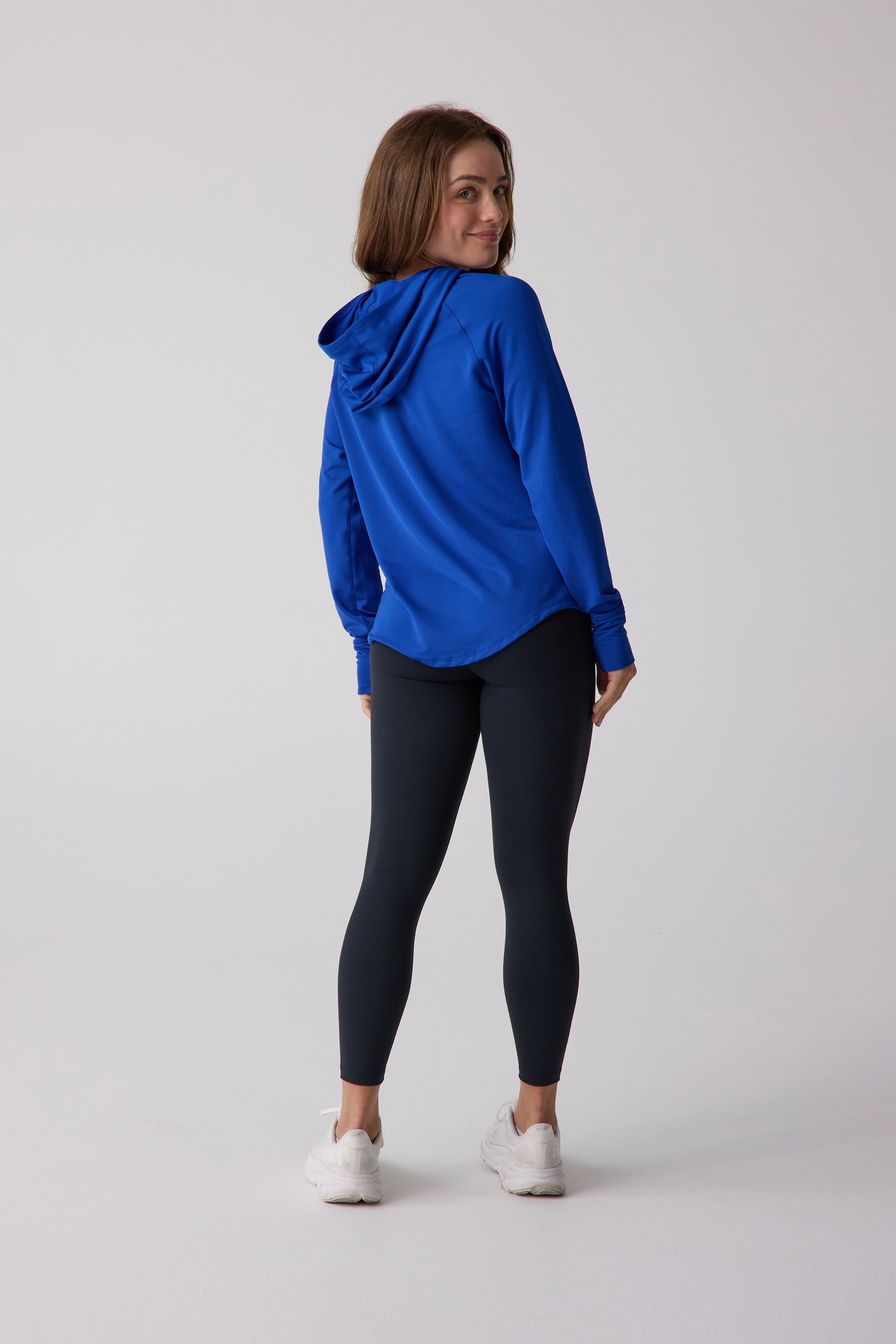 Blue women's layering hoodie by Centerline Athletics, back view with model smiling, paired with black leggings and white athletic shoes.