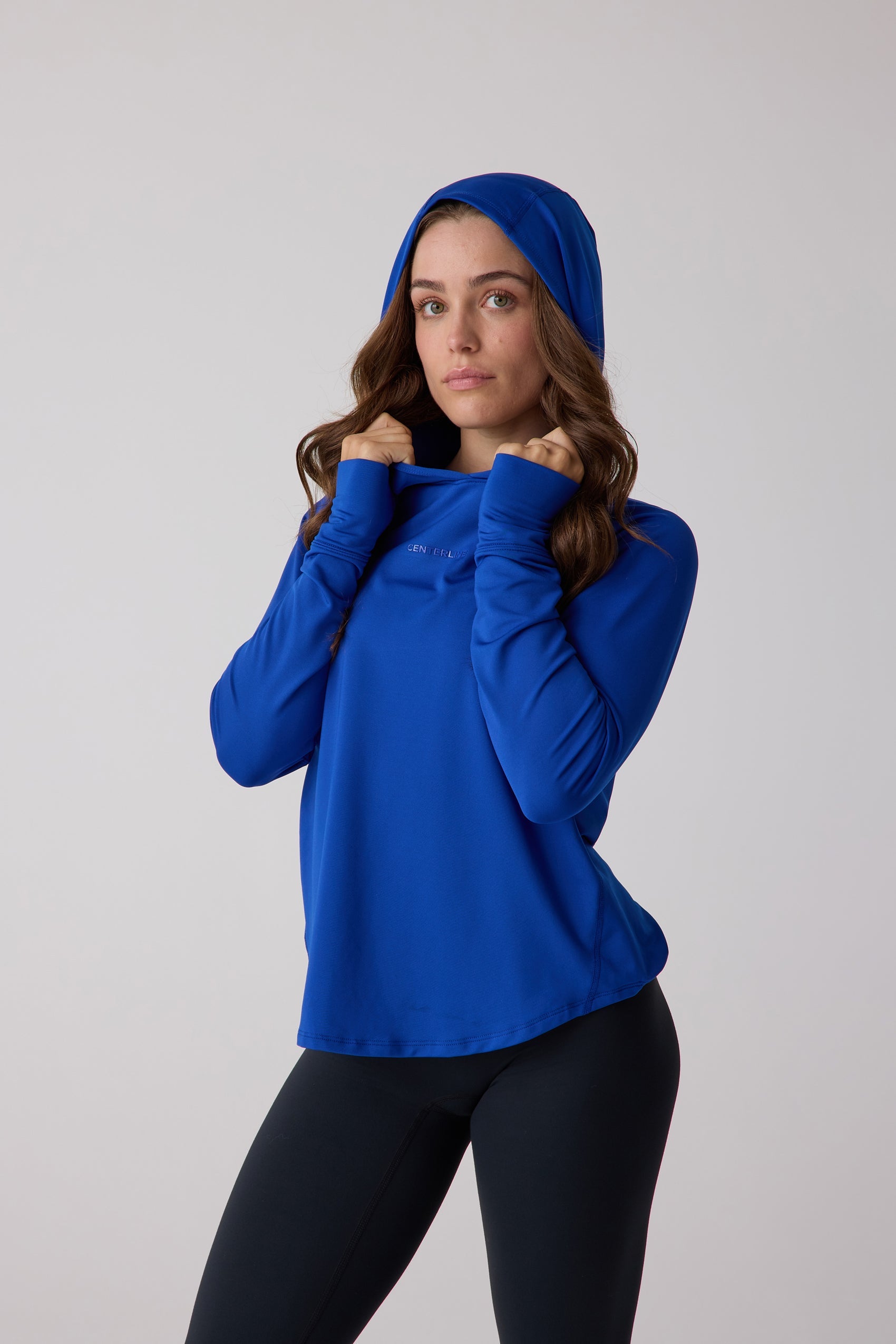 Blue women's layering hoodie by Centerline Athletics with long sleeves, thumbholes, and a subtle chest logo, styled over black leggings.