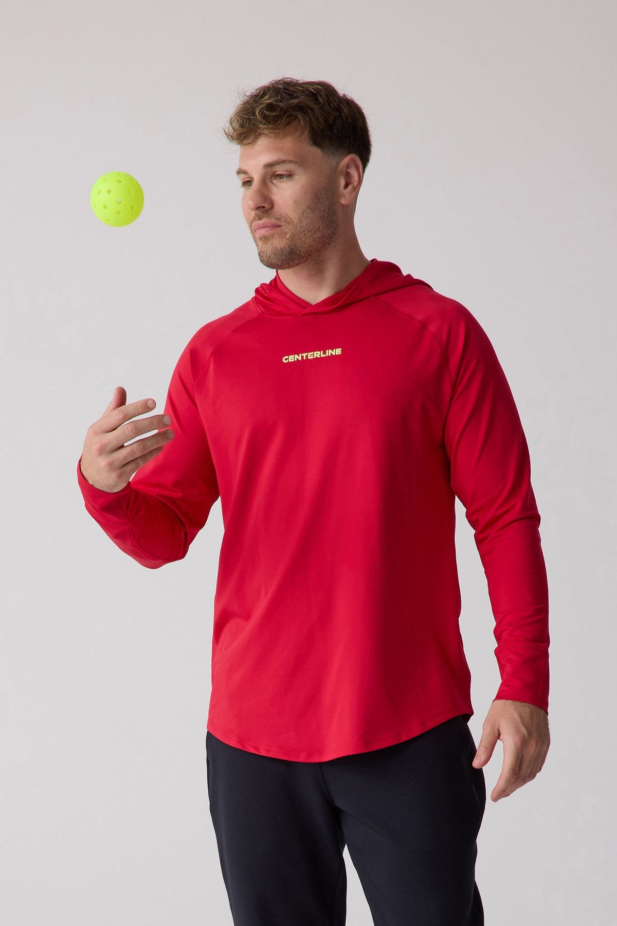 Red men's layering hoodie by Centerline with white logo, long sleeves, and hood, worn in lifestyle pose with yellow ball.