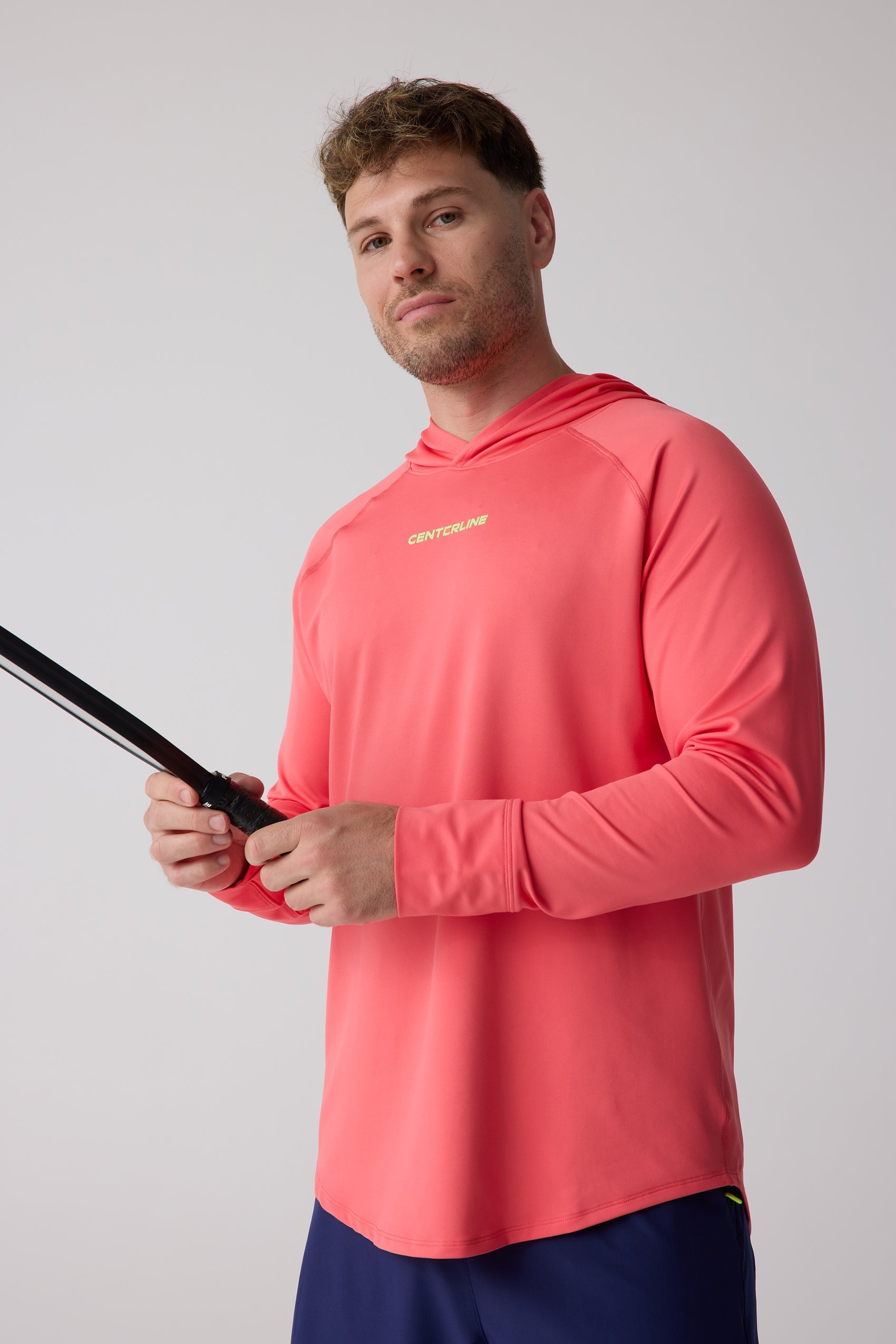 A coral men's layering hoodie by Centerline Athletics, featuring a neon yellow logo, raglan sleeves, and curved hem, worn by a man holding a black racket.