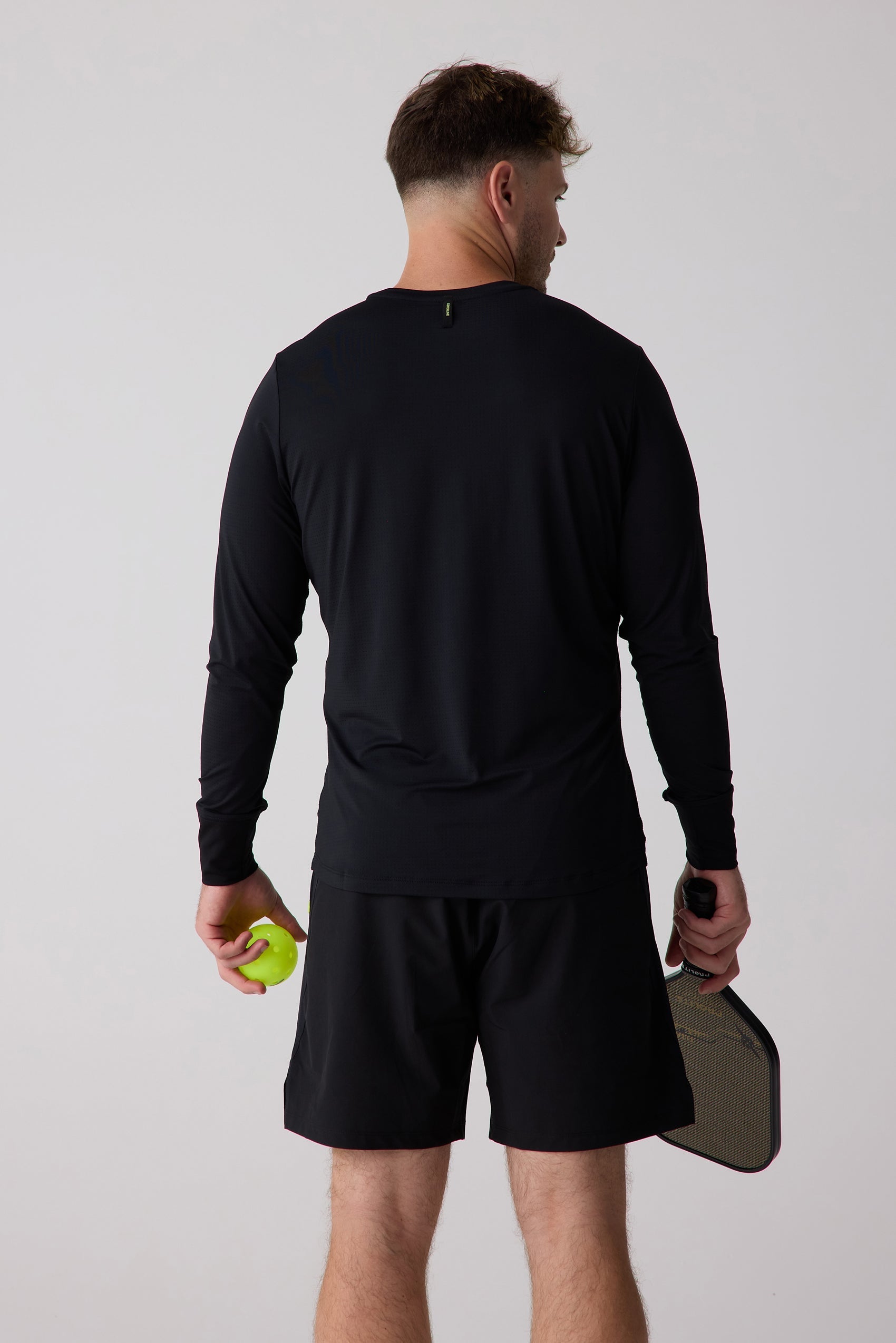 Black men's long sleeve shirt by Centerline Athletics, with subtle mesh texture and neon green collar tag, seen from the back holding a pickleball and paddle.
