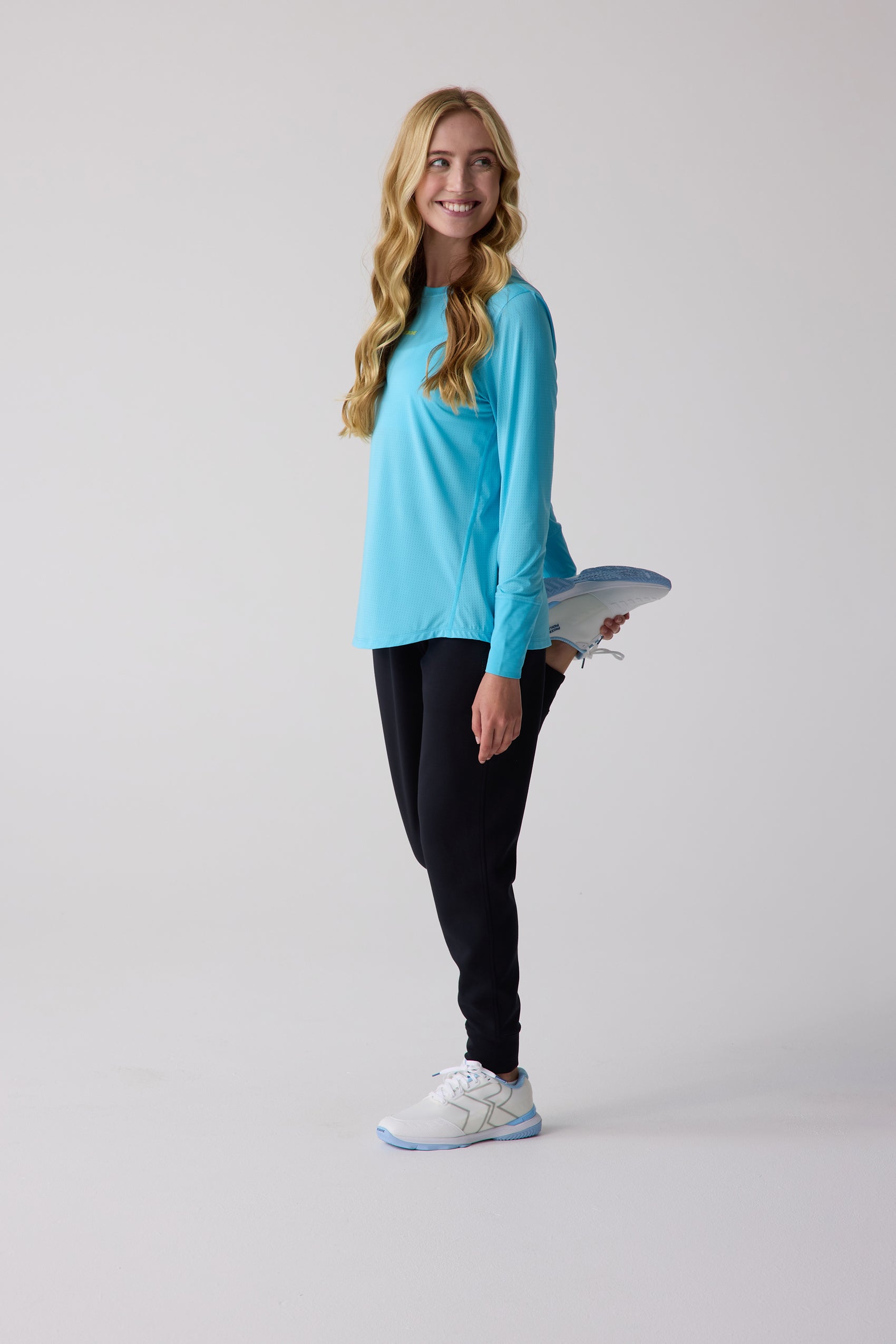 Women's turquoise blue perforated long sleeve shirt by Centerline Athletics, worn while stretching in black sweatpants and white athletic shoes.