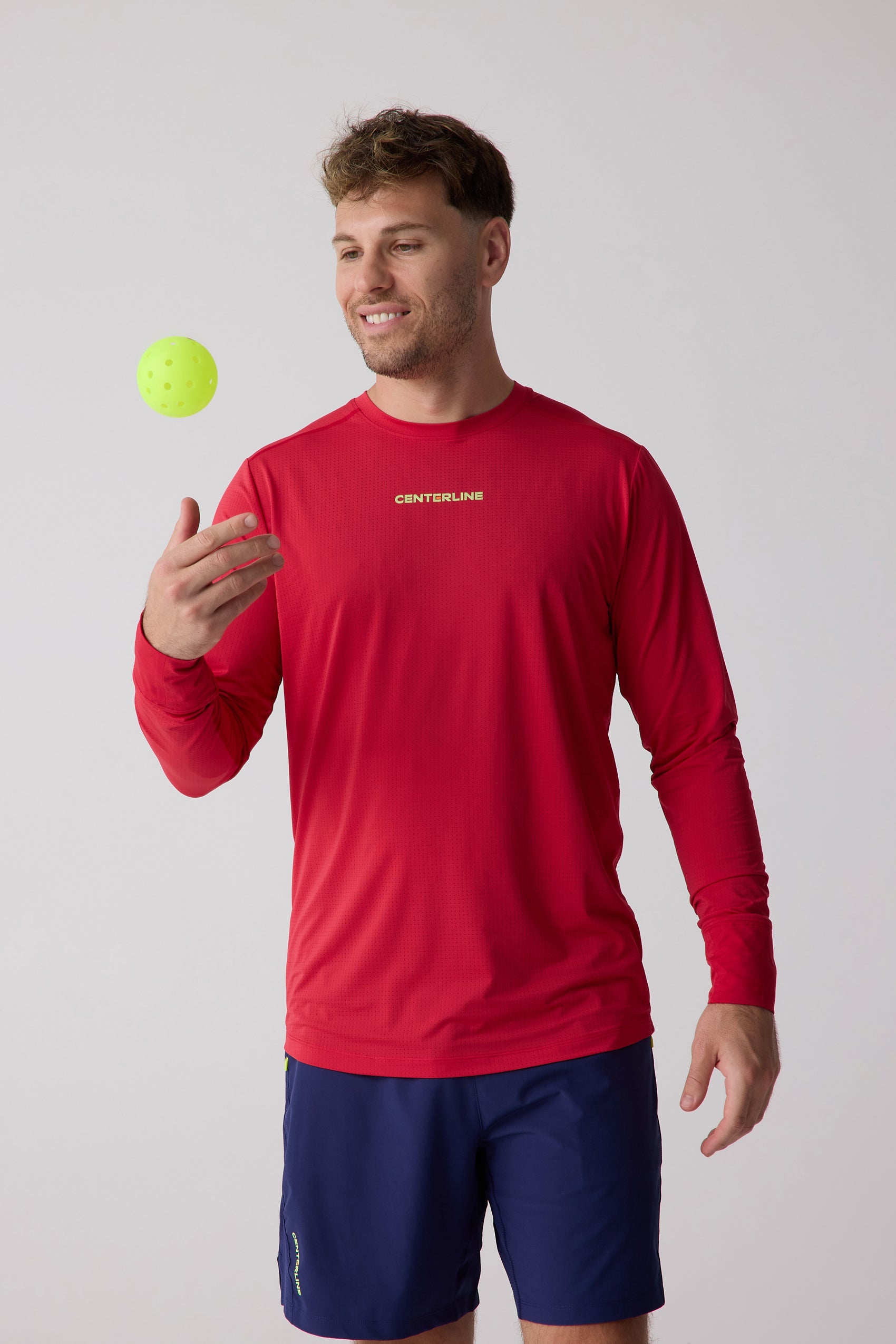 Red perforated men's long sleeve shirt by Centerline Athletics, with a yellow logo, worn by a man tossing a pickleball.