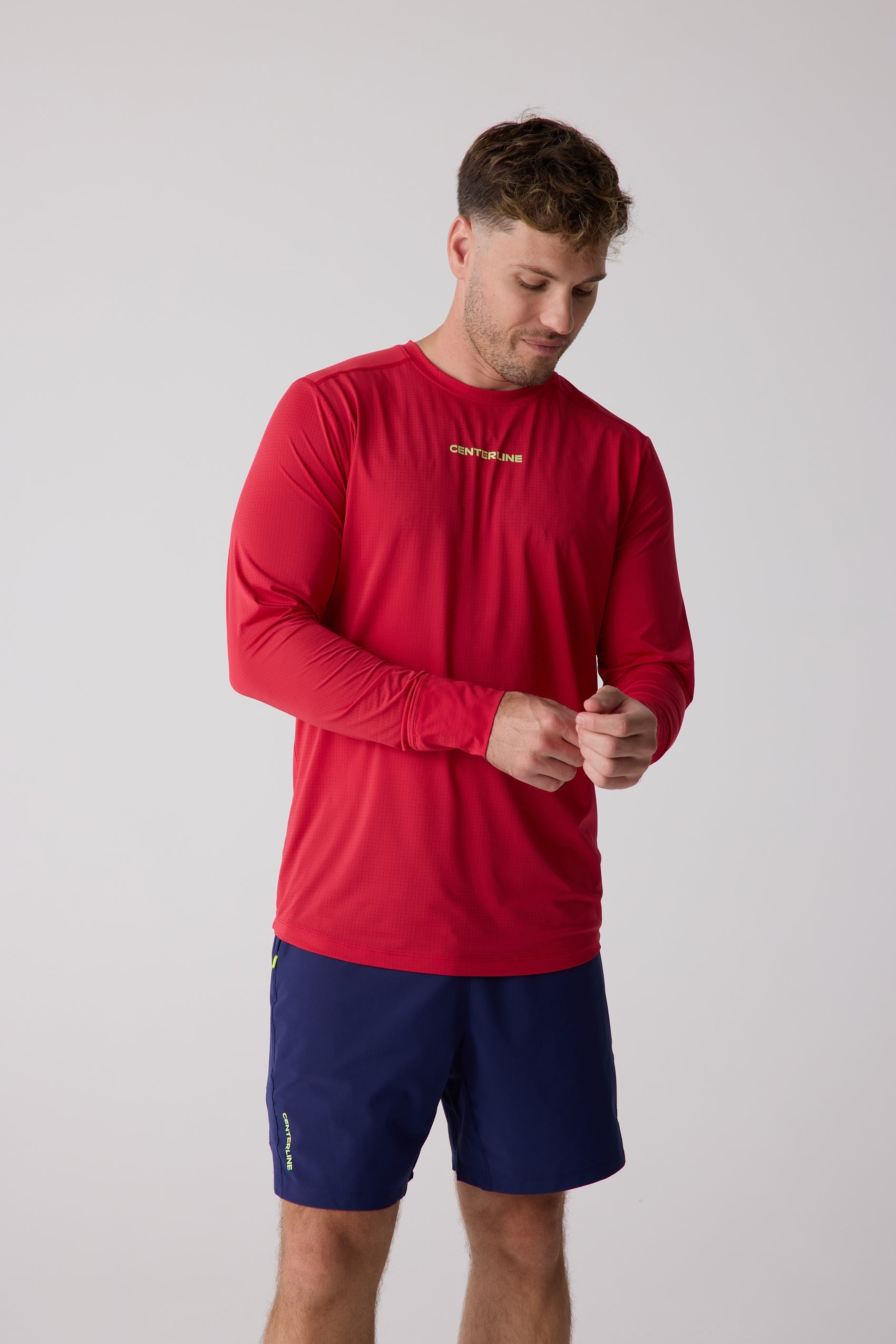 Red men's AirLite long sleeve shirt by Centerline Athletics with a perforated texture and yellow logo, worn by a man adjusting his cuff, alongside navy shorts.