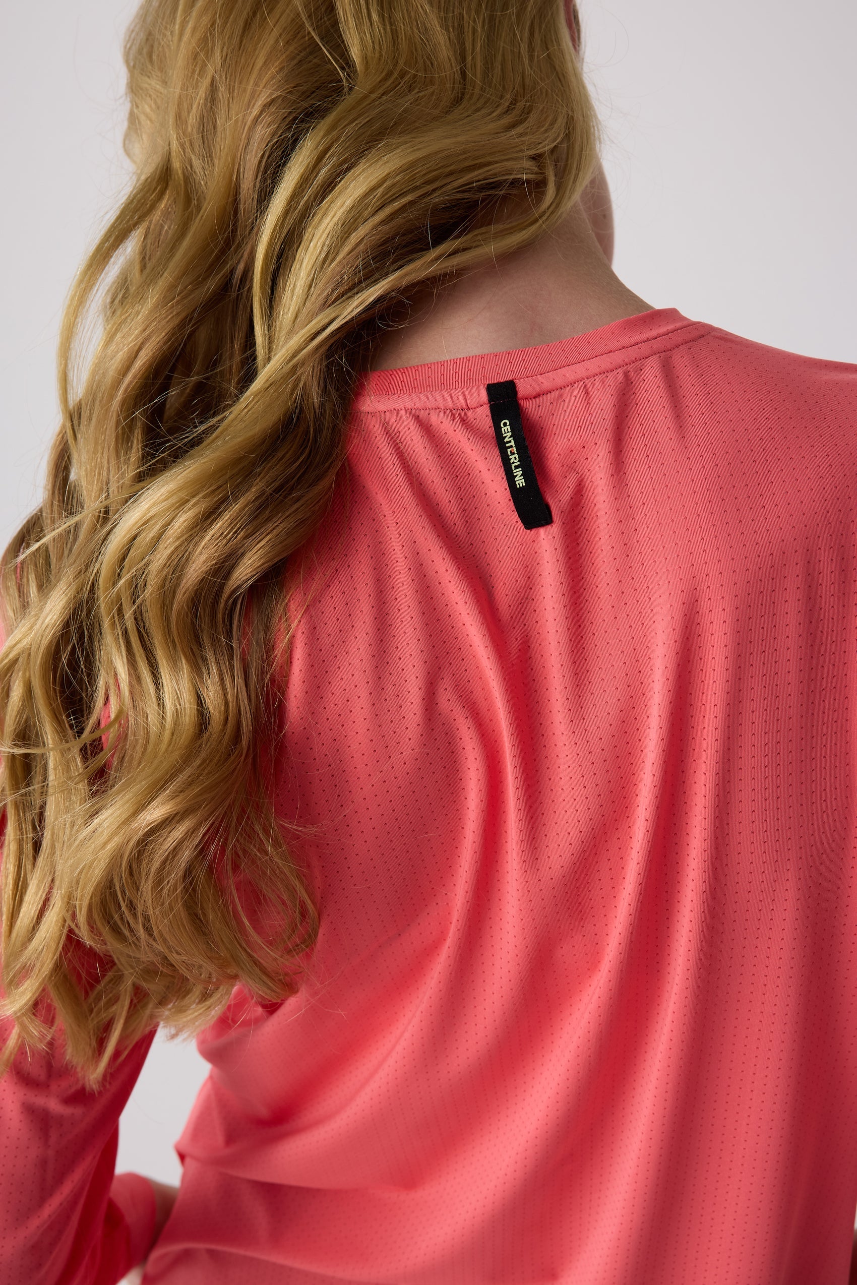 Coral women's long sleeve shirt by Centerline Athletics, with perforated fabric, a back tag, and long wavy blonde hair.
