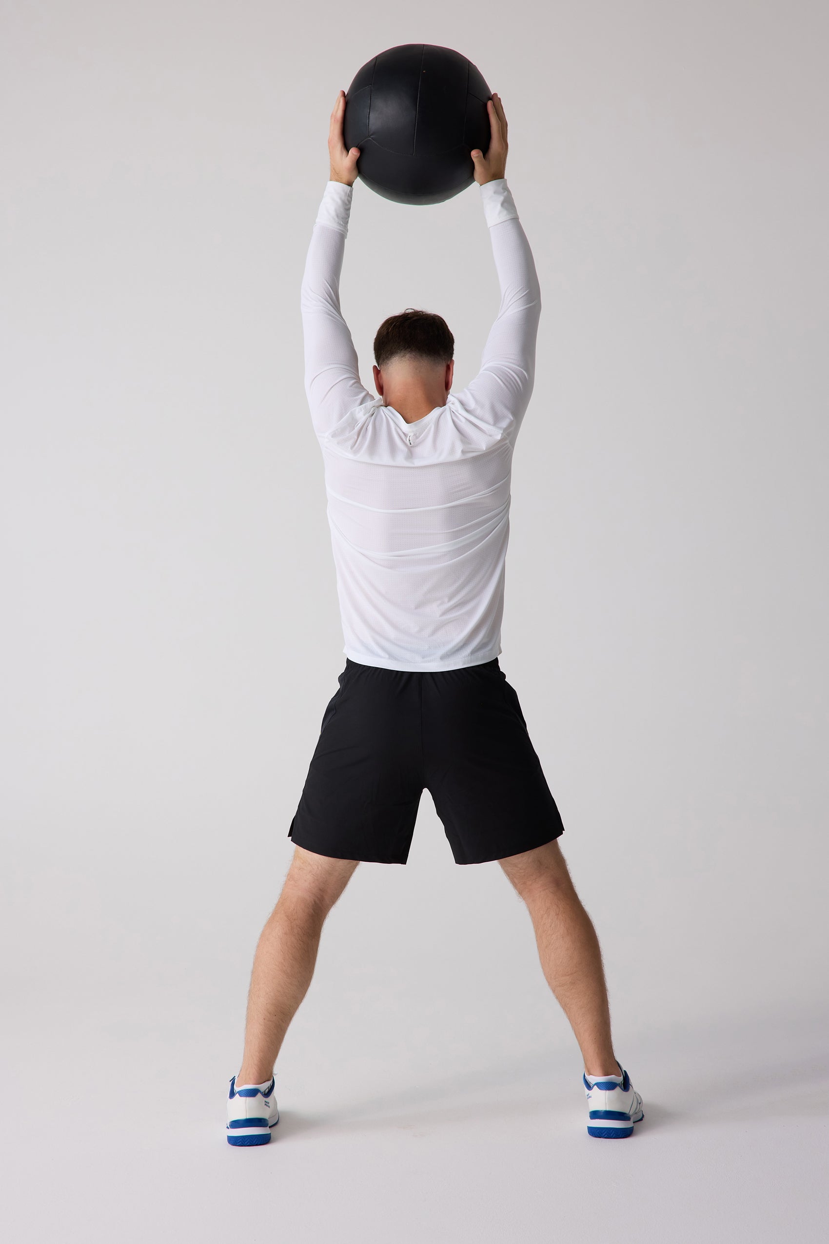 White Men's AirLite Long Sleeve by Centerline Athletics, mesh texture, back view, worn with black shorts and white sneakers, holding a black medicine ball overhead.