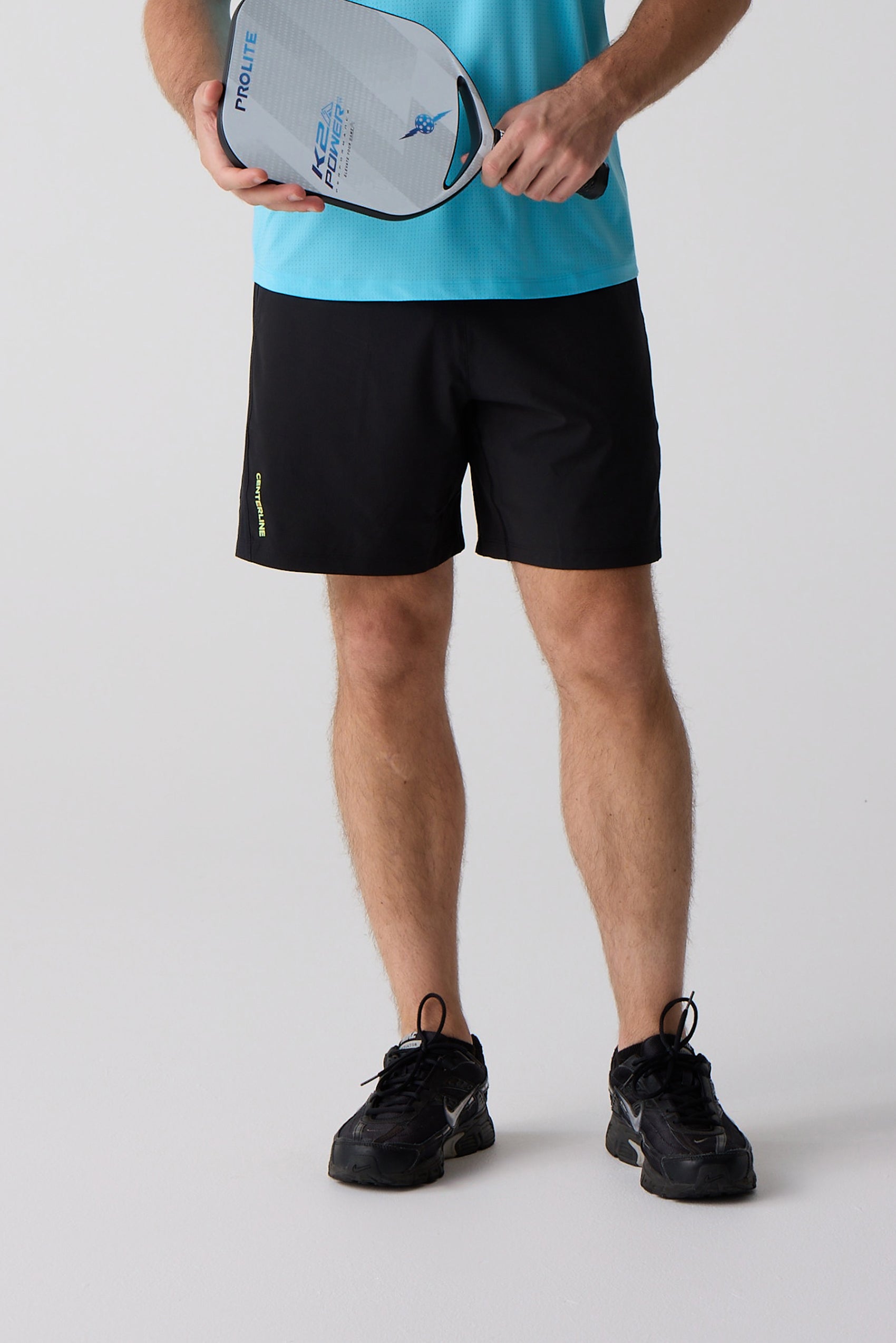 Black AirLite 7-inch unlined shorts by Centerline Athletics with neon yellow "CENTERLINE" text on the left leg, worn by a man holding a pickleball paddle.