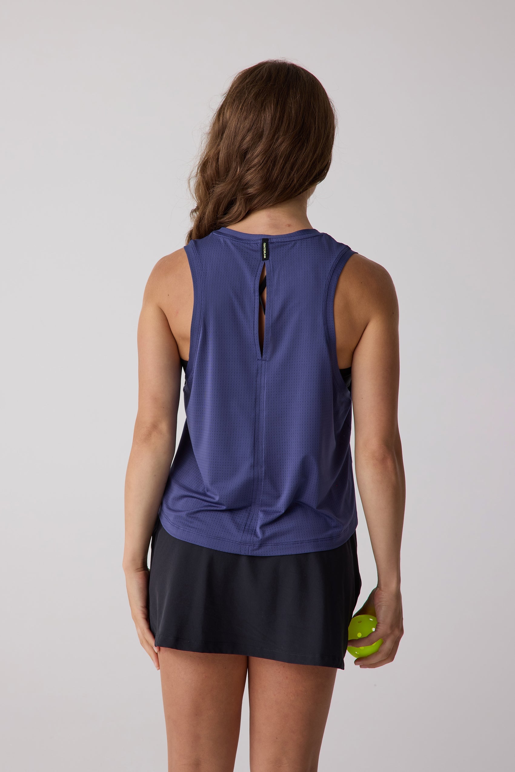 Perforated purple women's tank by Centerline Athletics, back view, with a keyhole cut-out, worn with a black skirt and holding a green pickleball.