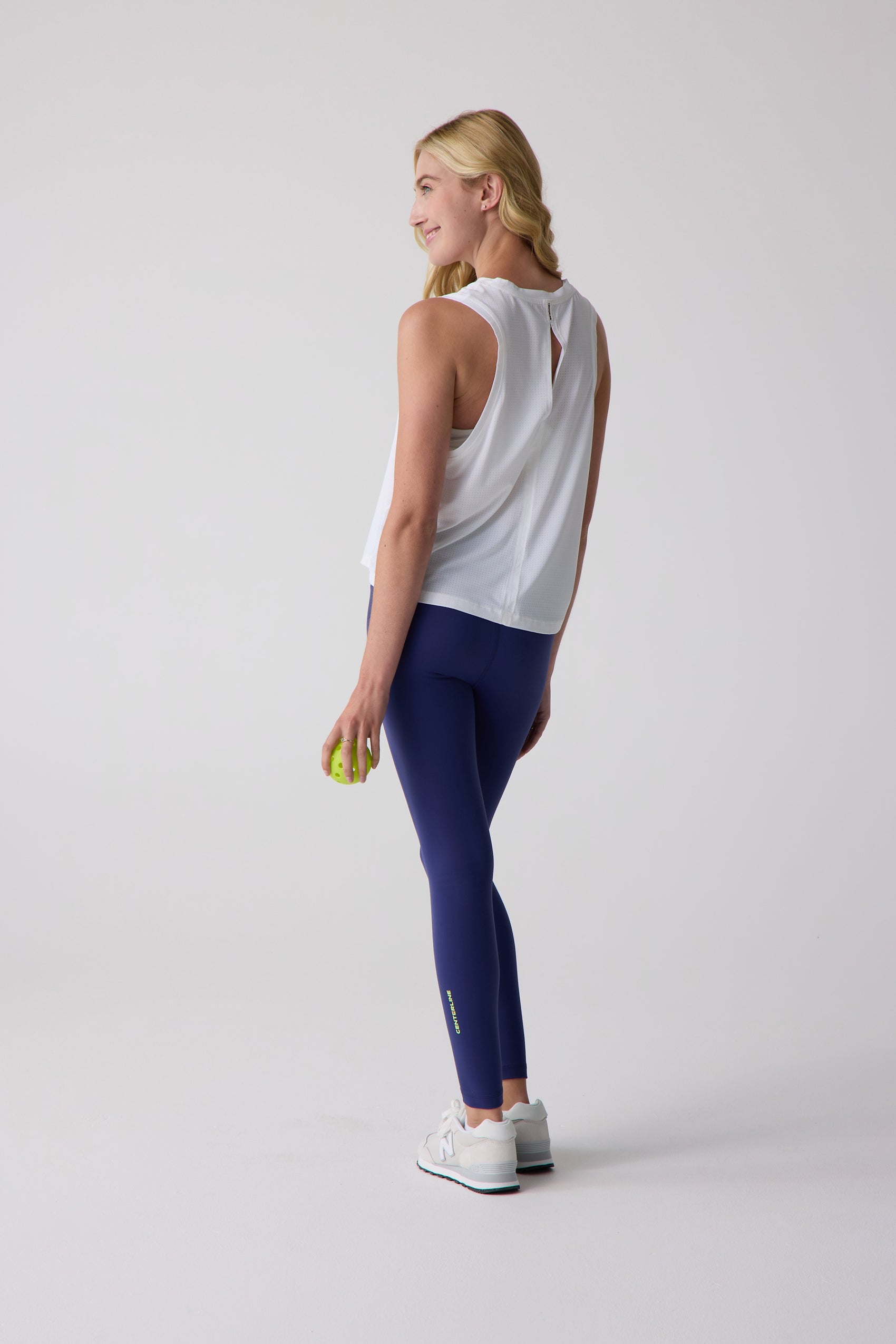White sleeveless AirLite tank by Centerline Athletics featuring a split back, worn by a woman in blue leggings and white sneakers.