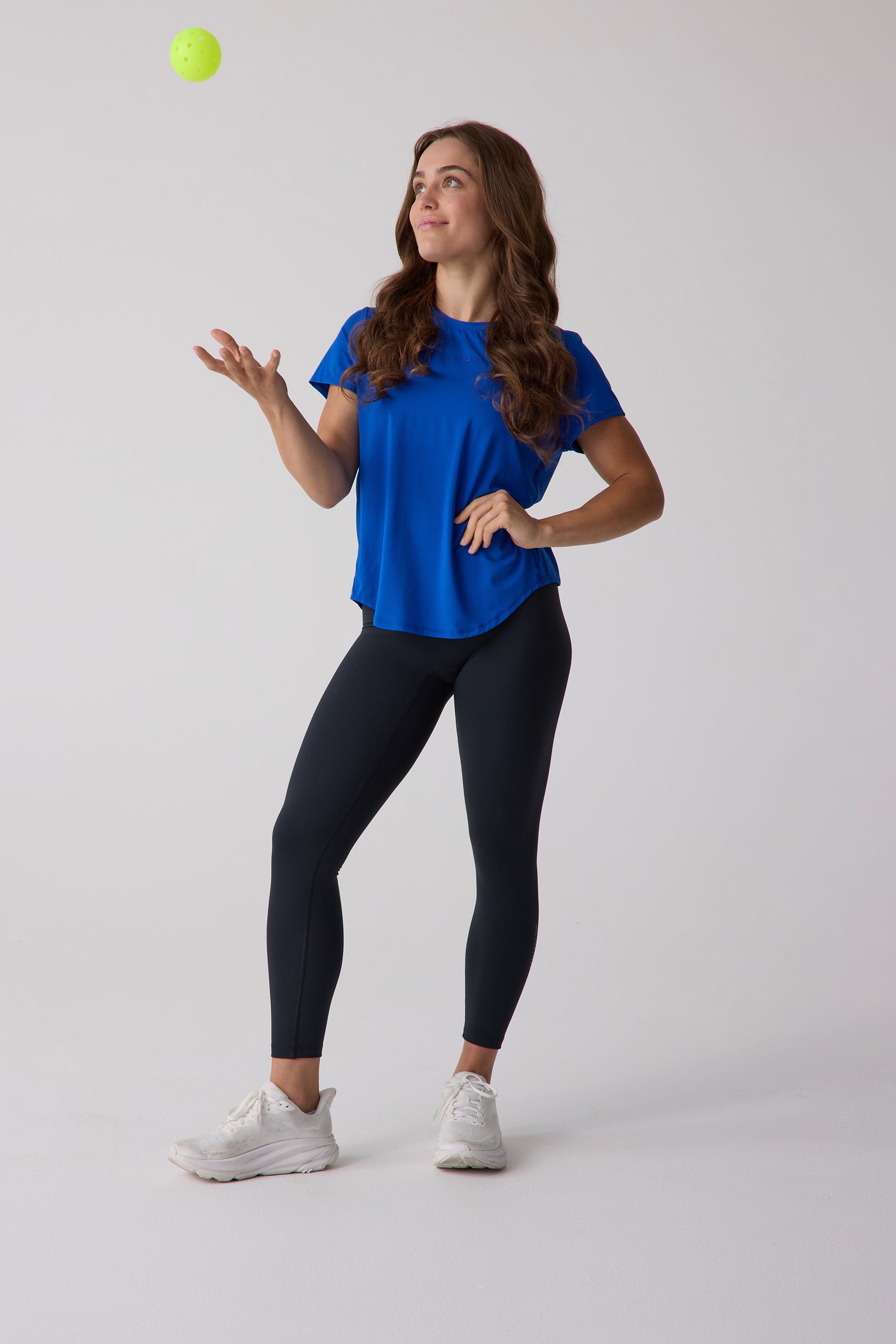Royal blue women's short sleeve tee by Centerline Athletics, with subtle texture, styled with black leggings and white sneakers, tossing a yellow pickleball.