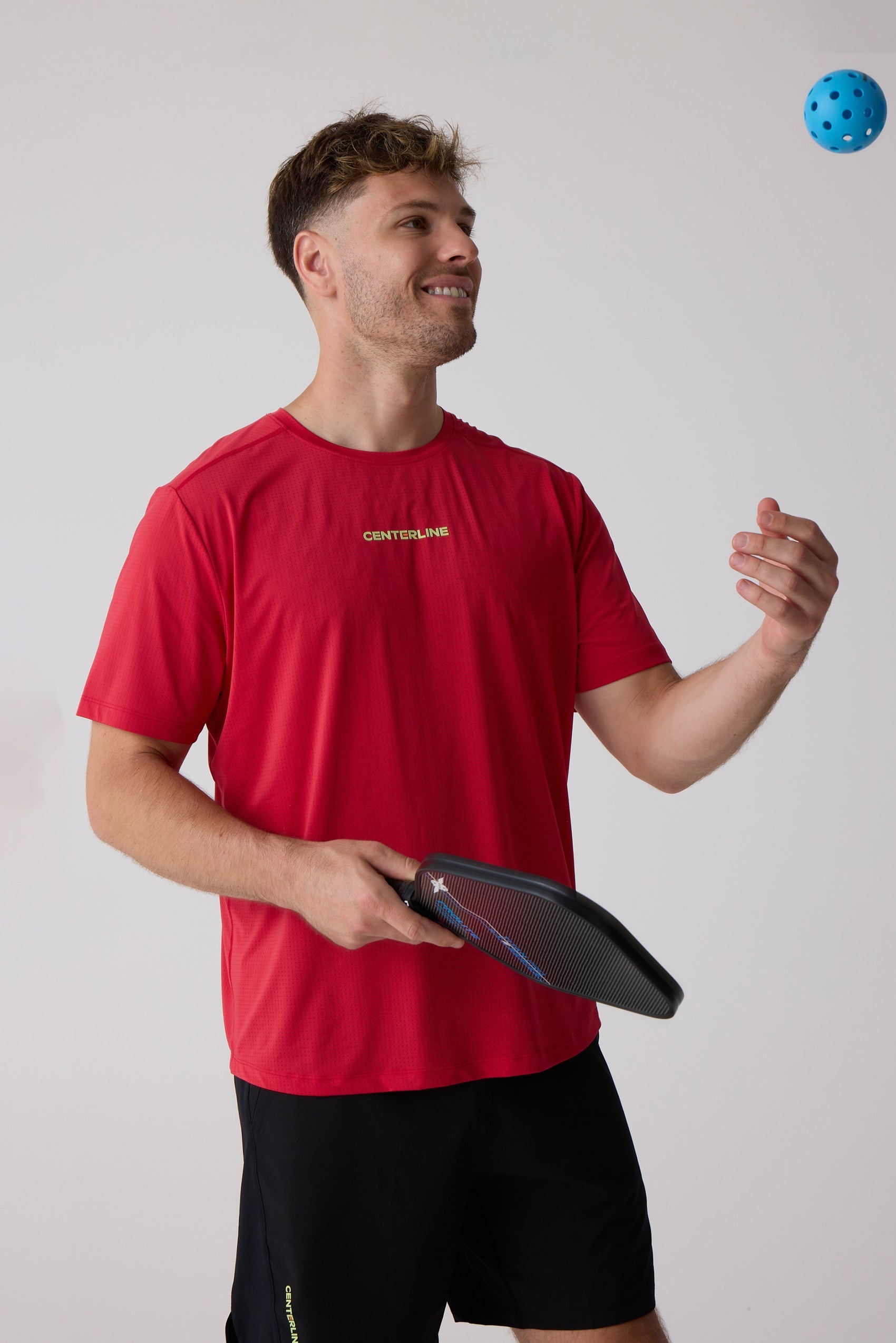 Red Men's AirLite Tee by Centerline Athletics, with a perforated texture and logo on the chest, worn by a man playing pickleball.
