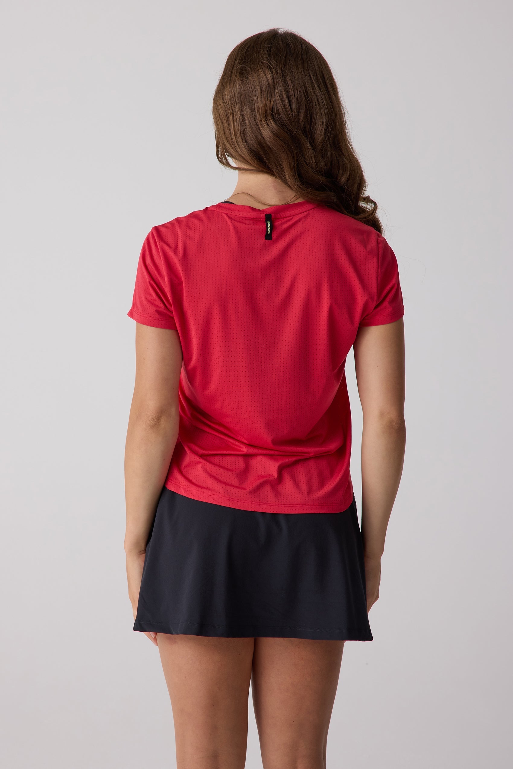 Red women's short sleeve AirLite tee by Centerline Athletics, featuring a perforated texture and small black back neck tag, shown from the back with a black skirt.