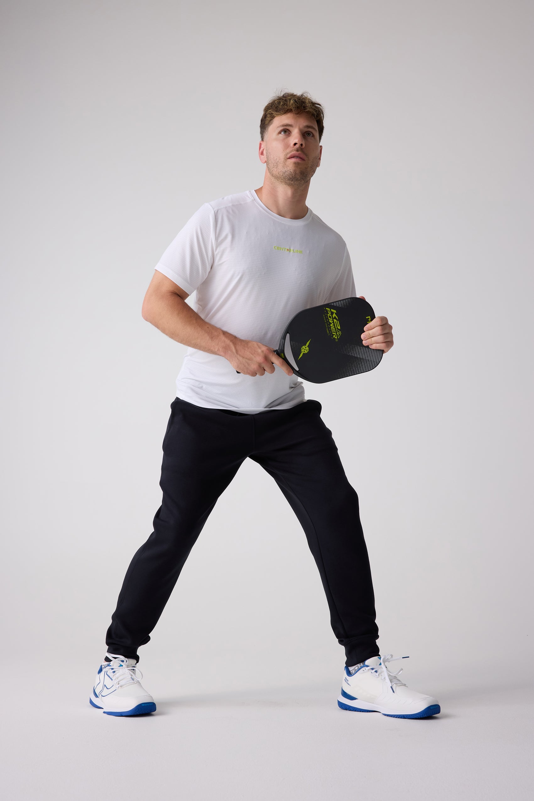 White Men's AirLite Tee by Centerline Athletics, with green 'CENTER-LINE' text and subtle textured pattern, worn by a man in black joggers holding a pickleball paddle.