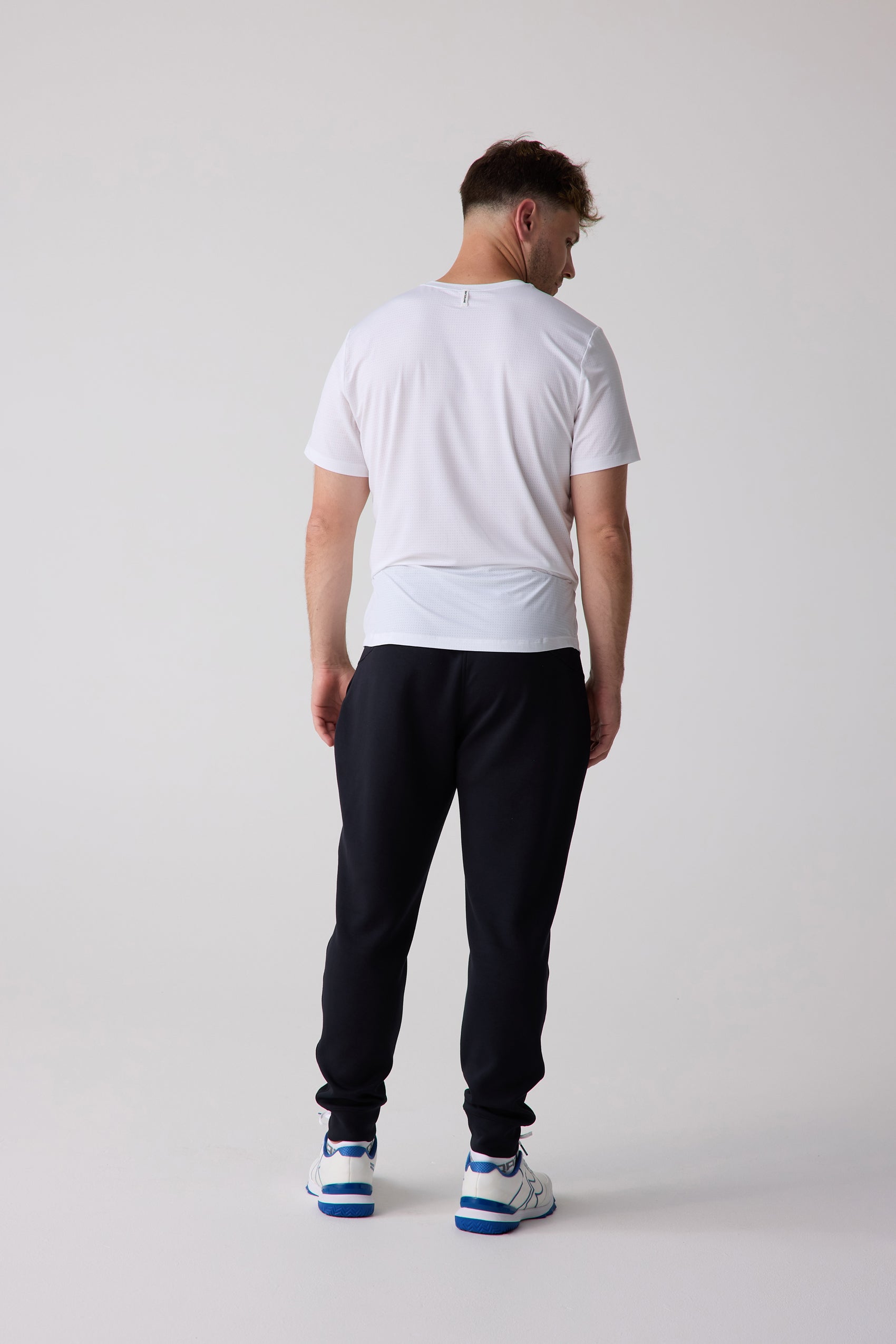 White, micro-perforated AirLite Tee by Centerline Athletics, back view, worn by a man with black joggers and blue/white athletic shoes.