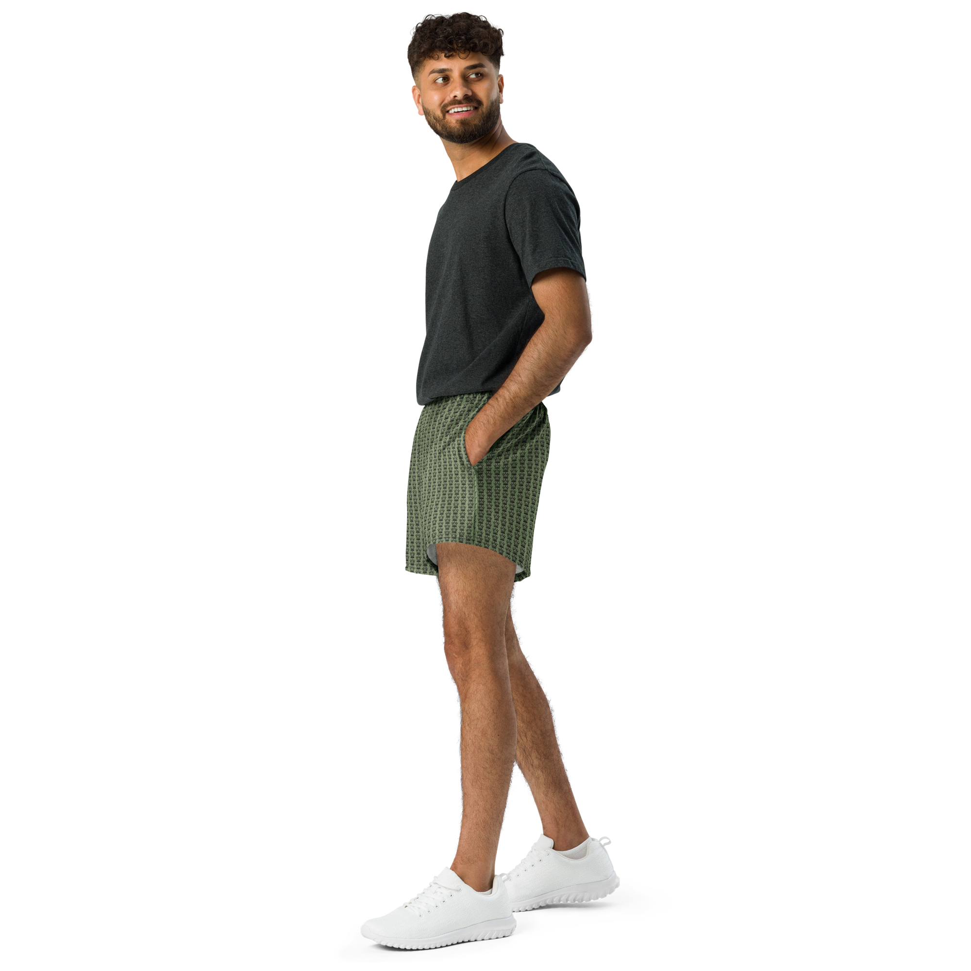 Man in a dark gray t-shirt, patterned olive shorts, and white sneakers. He is looking back with his hand in his pocket. He is standing against a black backdrop.