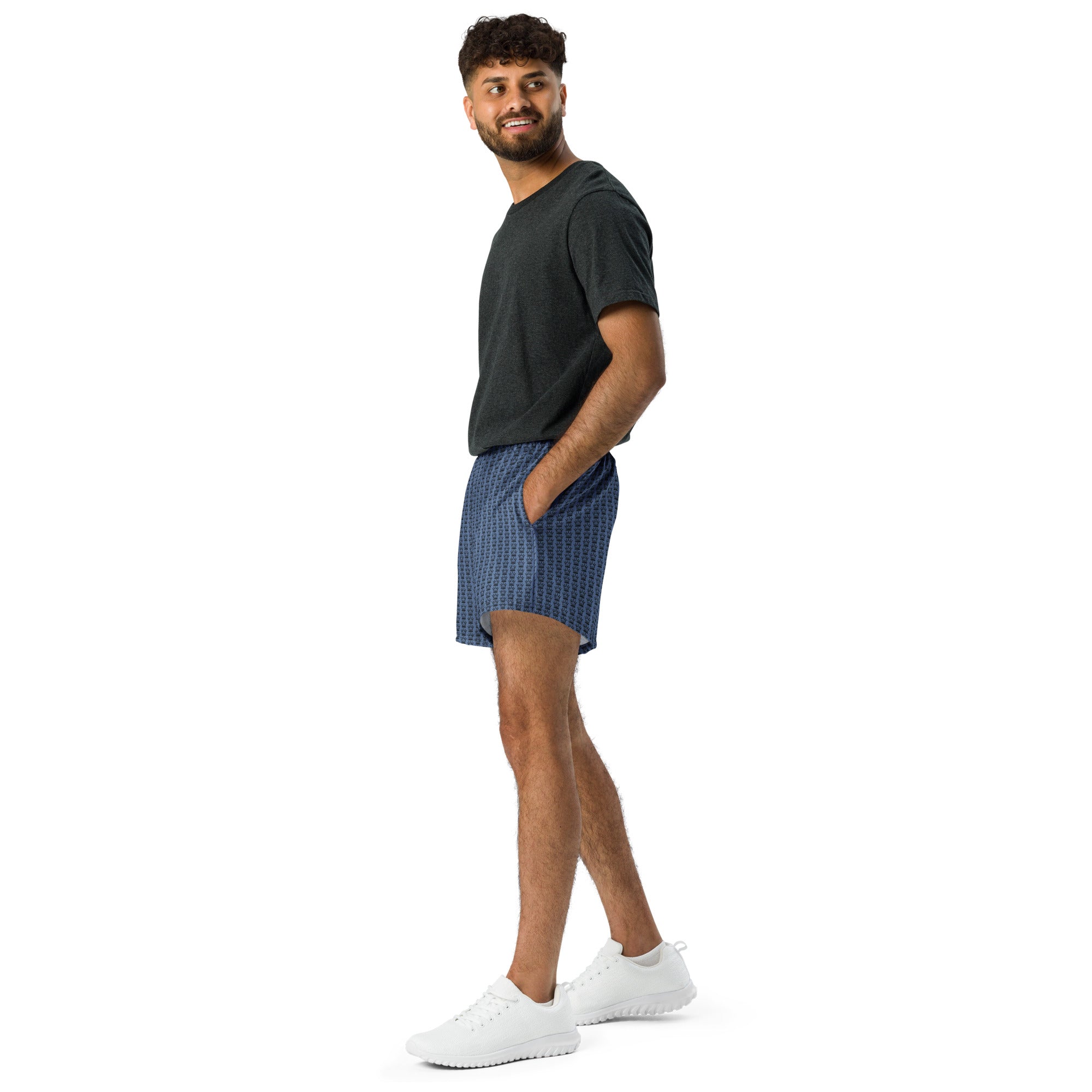 Man in a dark grey tee shirt and patterned blue shorts standing on a white background.