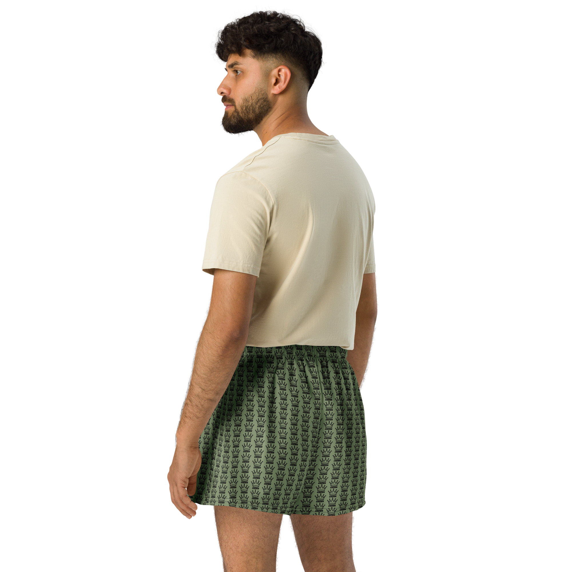 Man in tan t-shirt and green crown-patterned shorts, standing in profile against a black background.