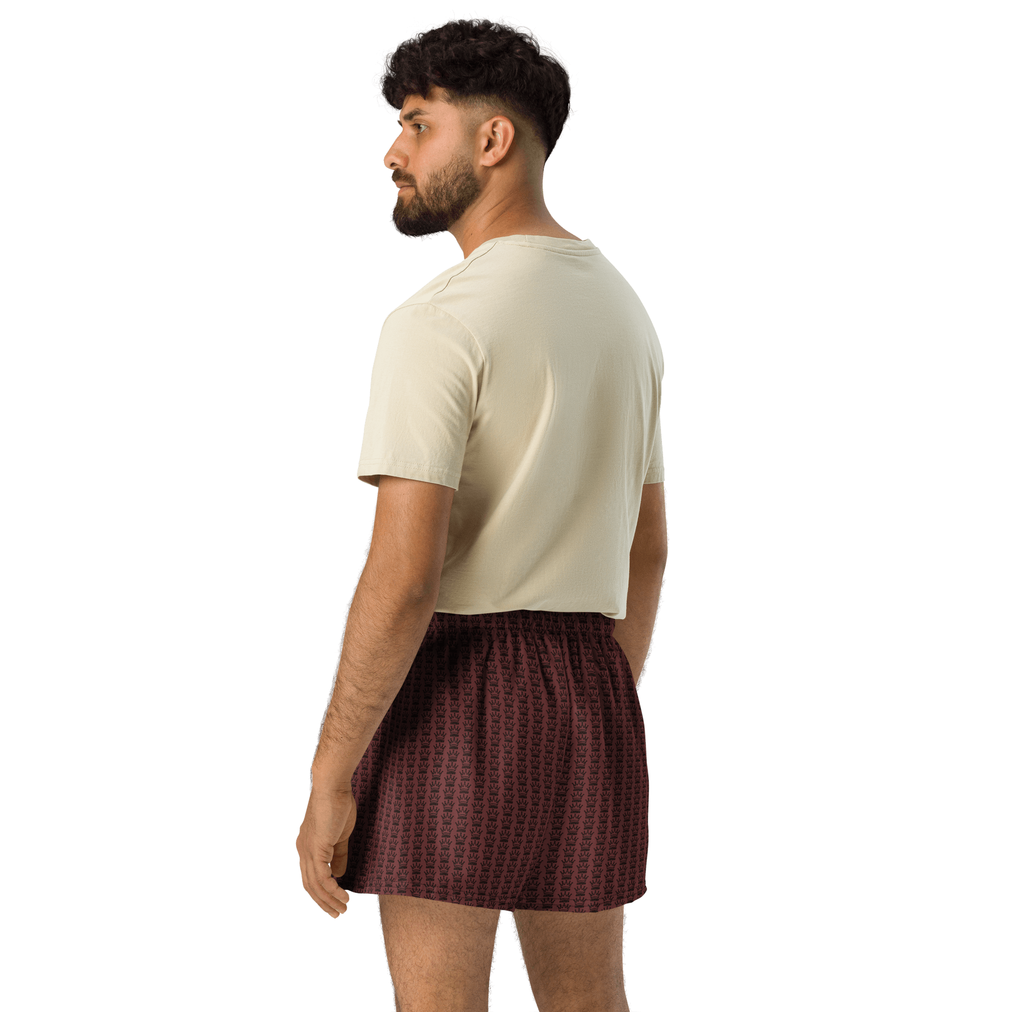 A man wearing a cream-colored t-shirt and burgundy boxer shorts with a crown pattern stands against a black background, turned to the side.