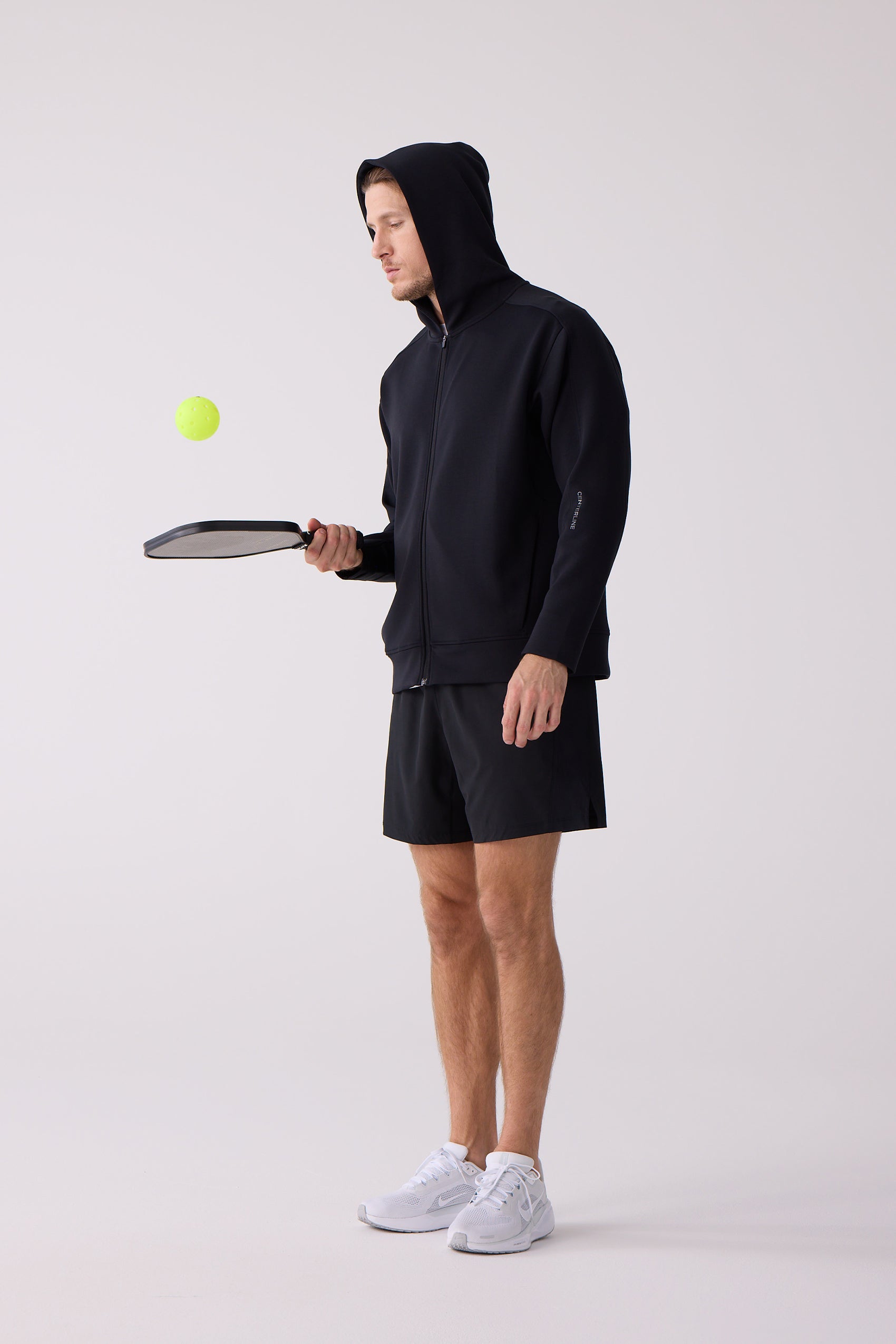 Black hooded full-zip sweatshirt by Centerline Athletics, with branding on sleeve, worn by a man playing pickleball.
