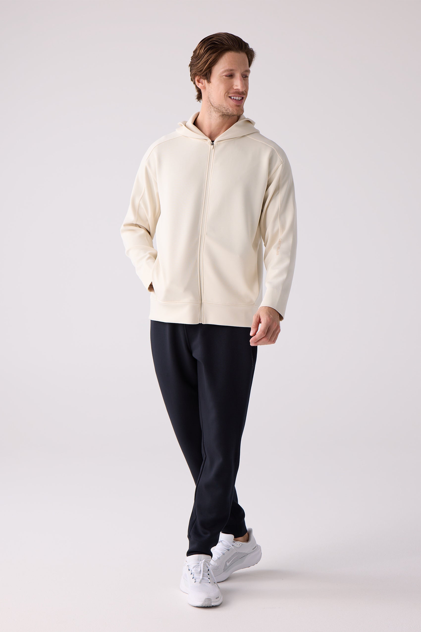 Light beige full-zip hoodie by Centerline Athletics with 'CENTERLINE' text on sleeve, worn by a smiling man with dark sweatpants and white sneakers.