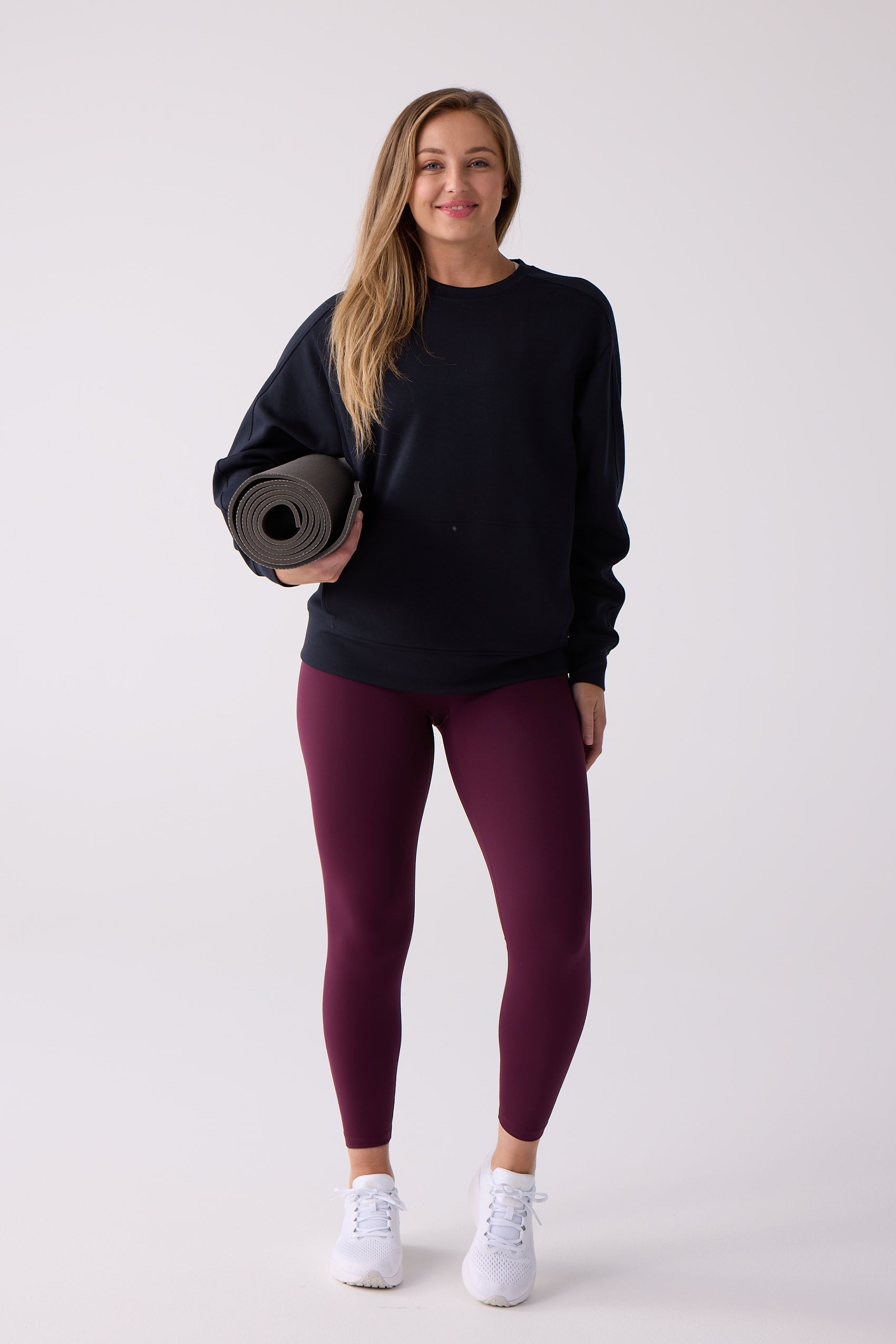 Black Women's Crew Sweatshirt by Centerline Athletics, worn with maroon leggings, white sneakers, and holding a grey yoga mat.