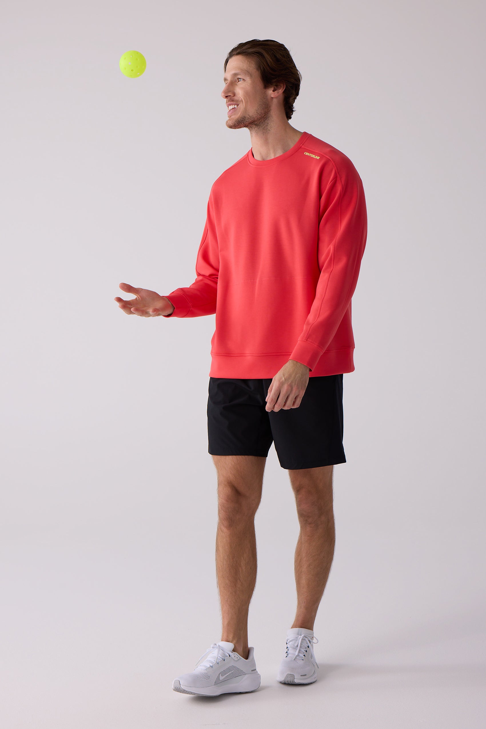 Red men's crew sweatshirt by Centerline Athletics, with yellow branding on the shoulder, worn by a man in black shorts and white sneakers tossing a pickleball.
