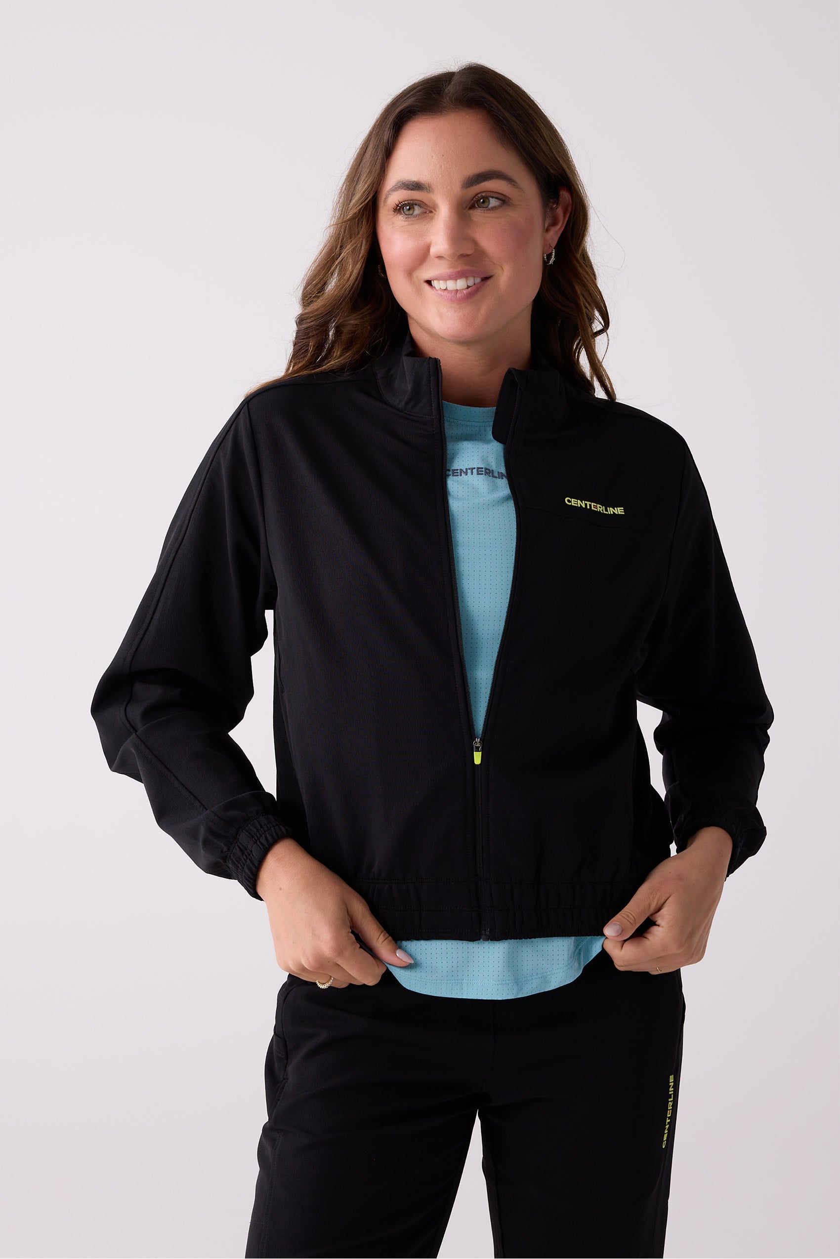 Black Women's Courtside Track Jacket by Centerline Athletics, featuring a neon yellow logo and zipper, worn over a light blue textured top.