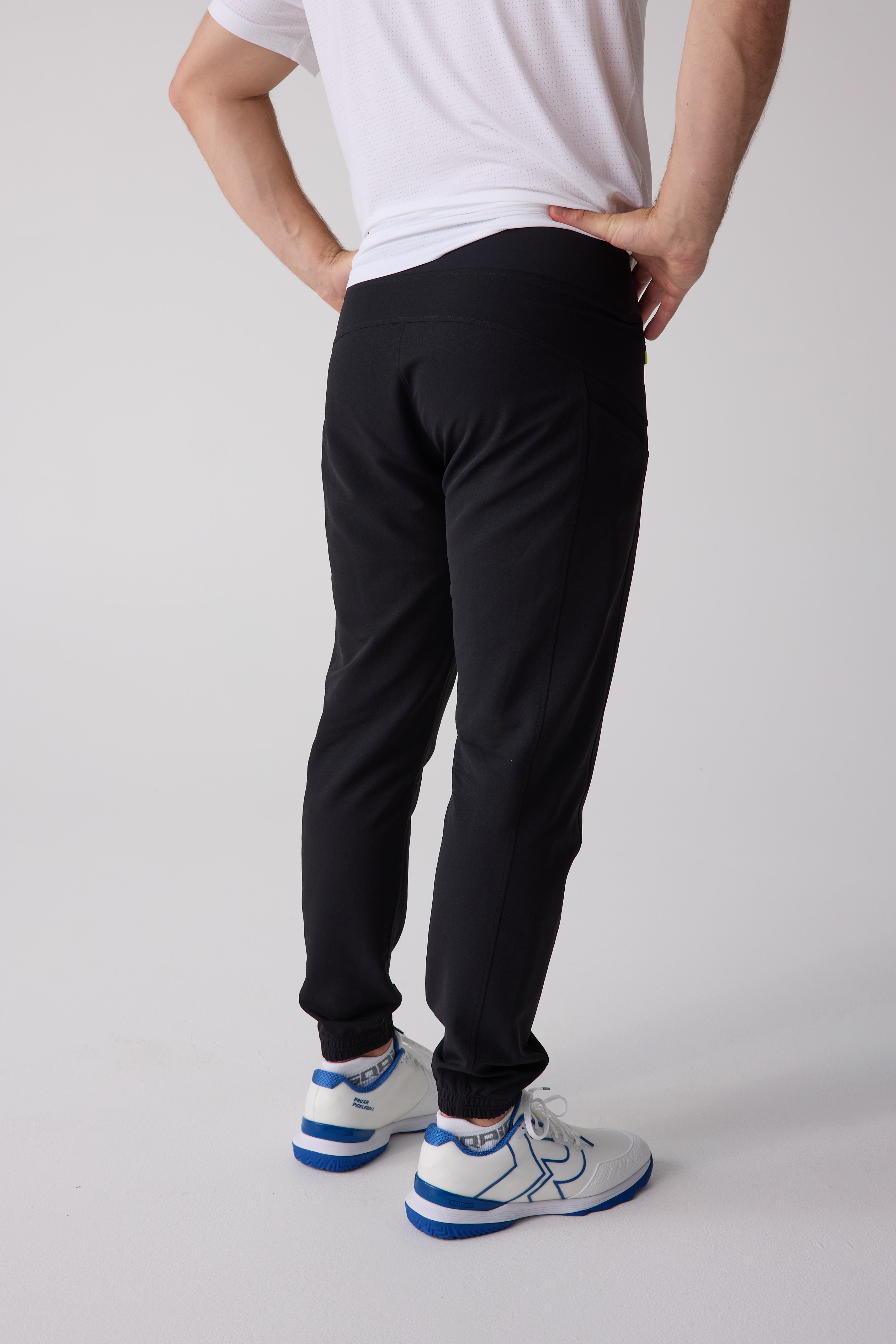 Black Men's Courtside Track Pants by Centerline Athletics, rear-side view, worn with a white perforated t-shirt and white and blue athletic shoes.