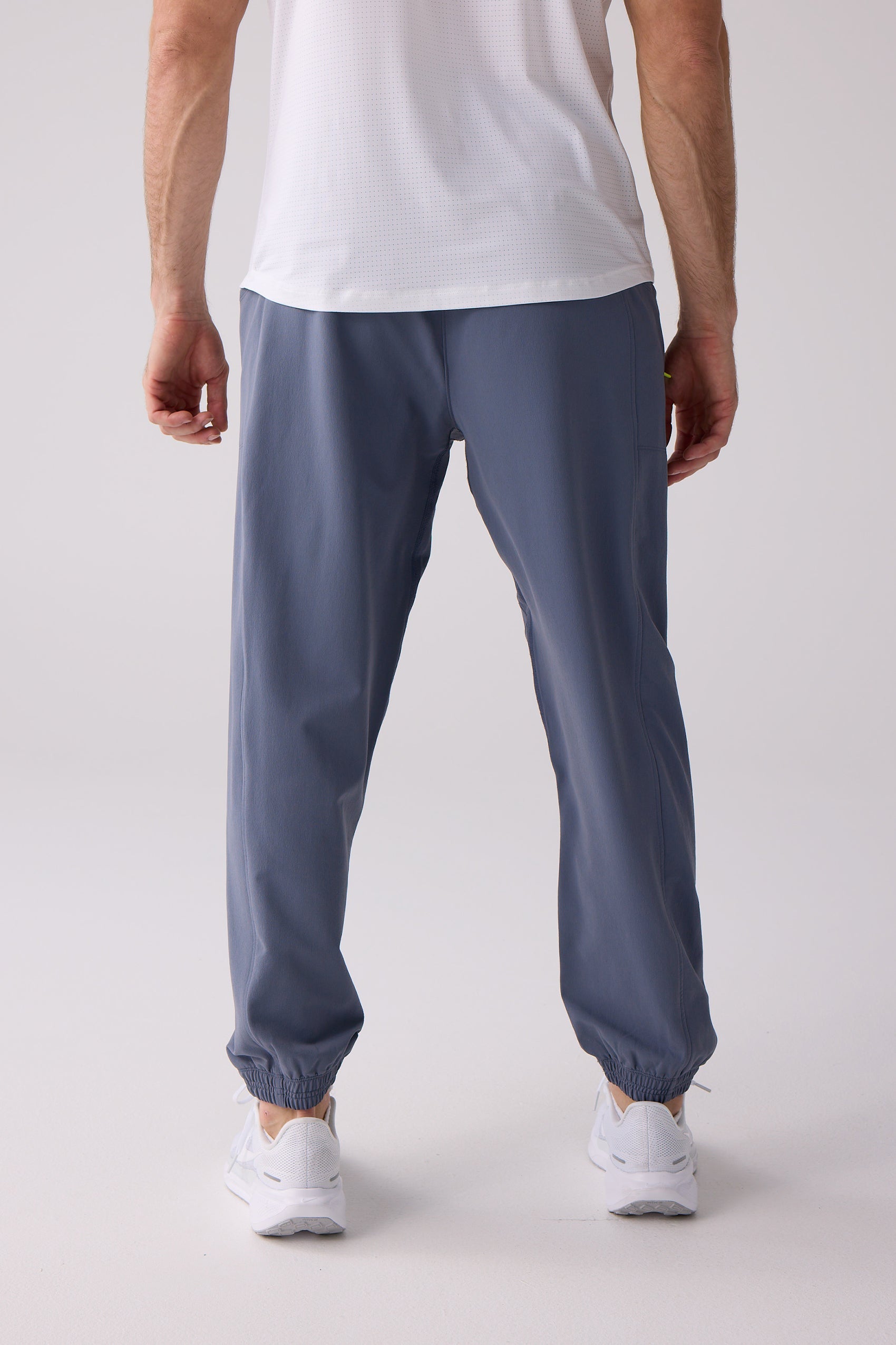 Men's slate blue track pants by Centerline Athletics, with elasticized cuffs, a relaxed fit, worn with a white perforated shirt and white sneakers, shown from the back.