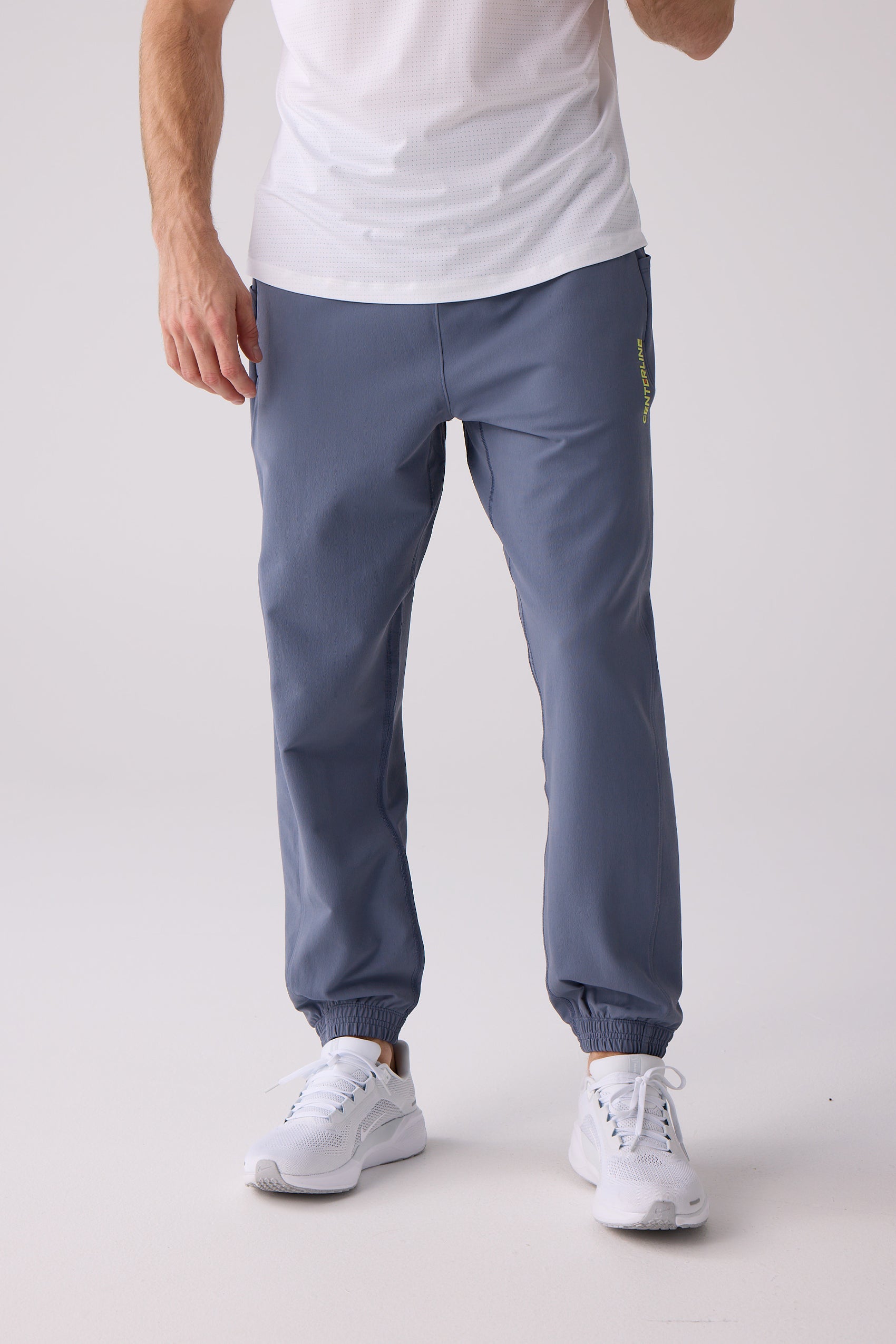 Men's slate blue track pants by Centerline Athletics, with a vertical yellow 'CENTERLINE' logo and elasticated cuffs, worn with a white t-shirt and sneakers.
