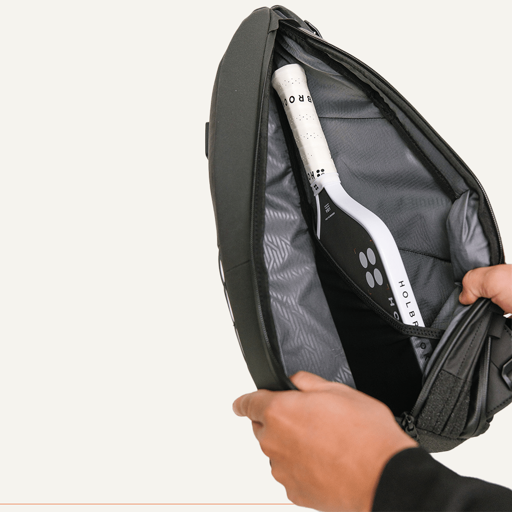 Dark gray pickleball sling from FORWRD, open with a black and white pickleball paddle inside, held by a hand.