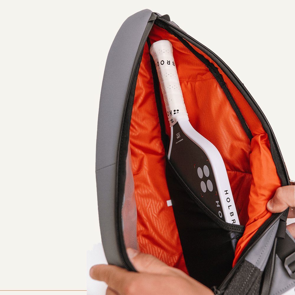 Grey fabric pickleball sling by FORWRD with an open orange interior holding a pickleball paddle.
