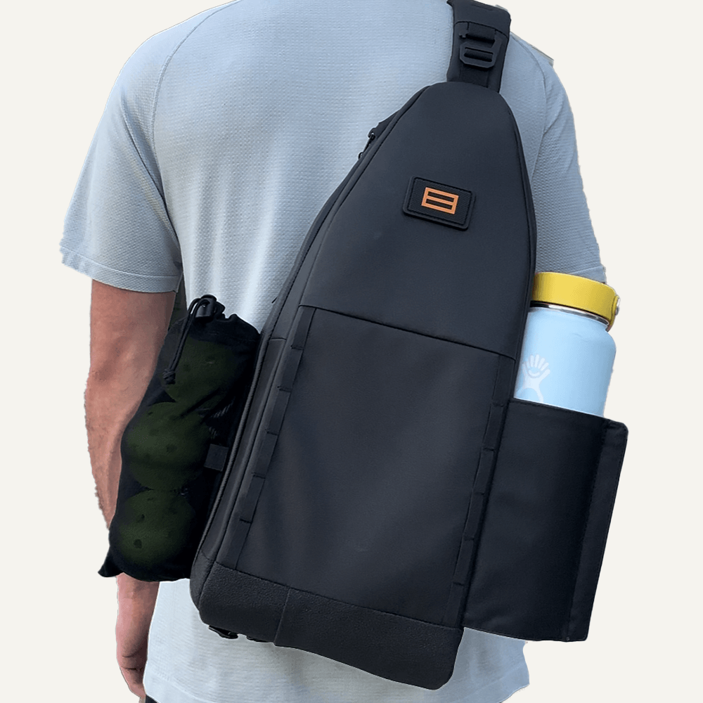 Person wearing a black sling backpack with a water bottle in a side pocket and a mesh bag with tennis balls.