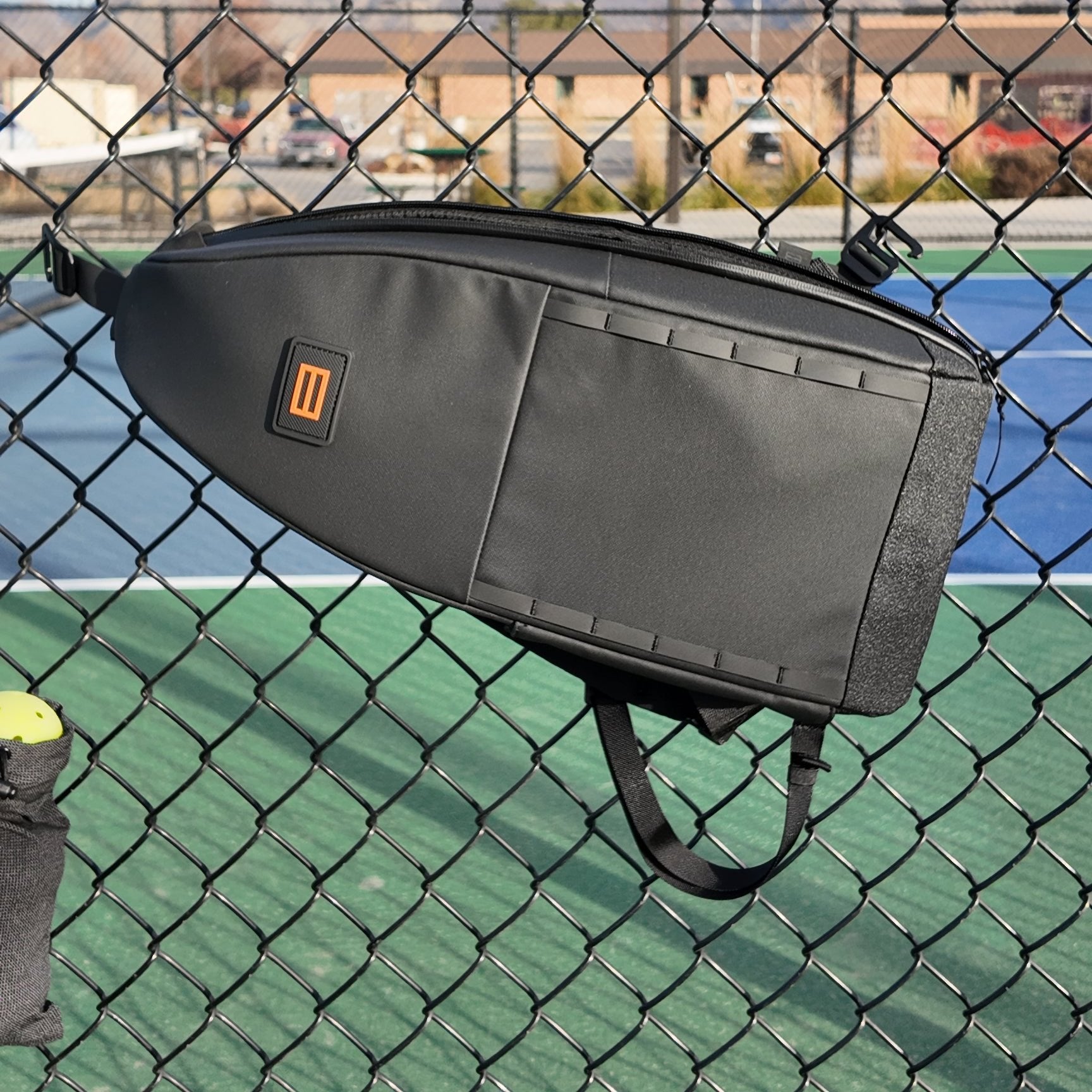 Black Cross Court Pickleball Sling by FORWRD, with an orange logo and textured panels, hanging on a fence over a court with a yellow pickleball.