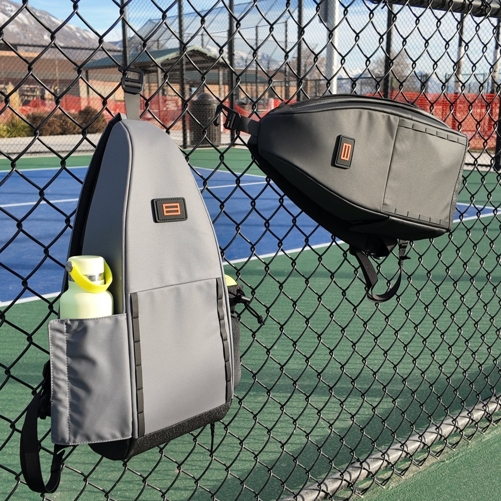 Two bags hung on a chain-link fence, one grey with a water bottle in the side pocket, and one dark grey.
