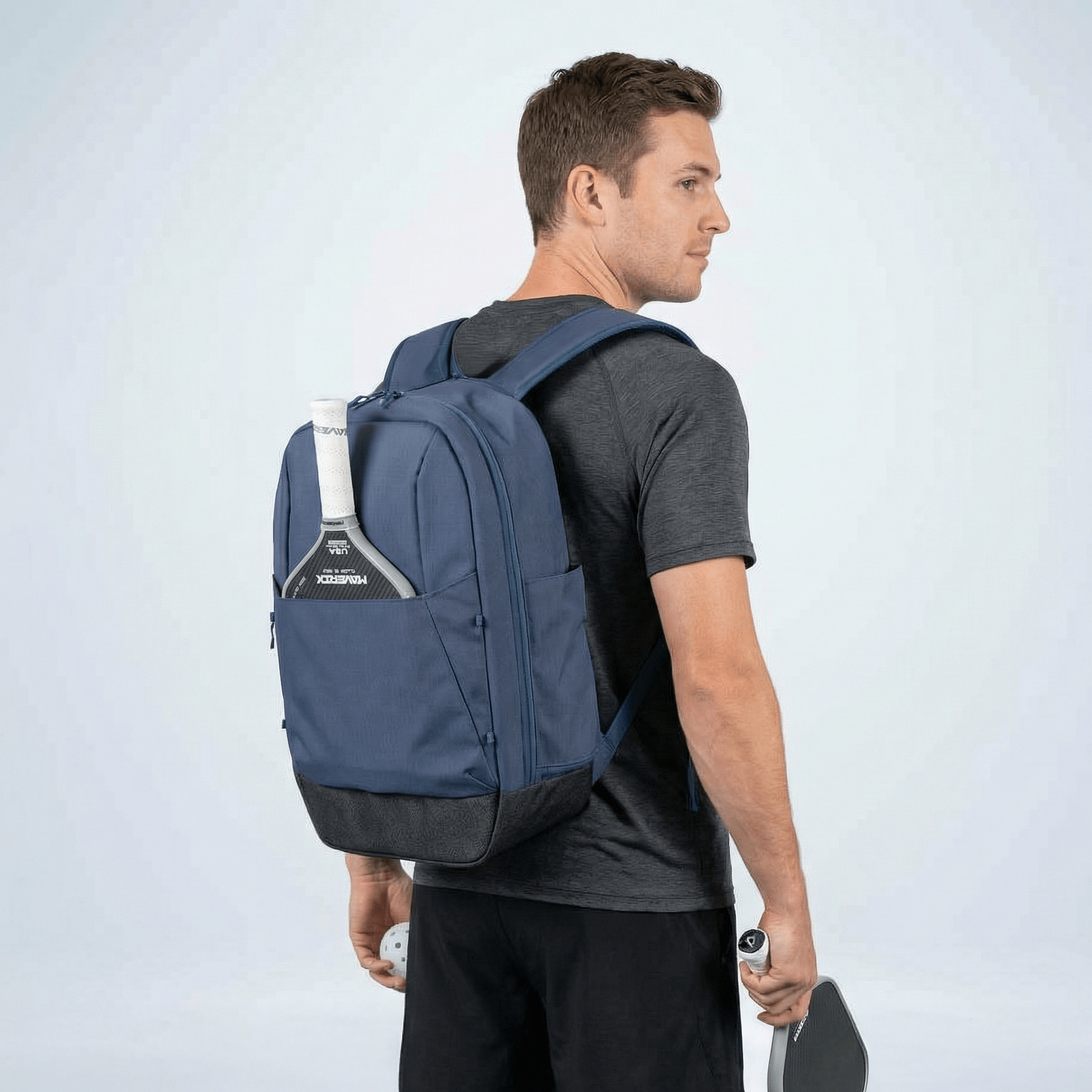 Navy blue and dark grey pickleball backpack by FORWRD worn by a man from behind, with a paddle in its side pocket.