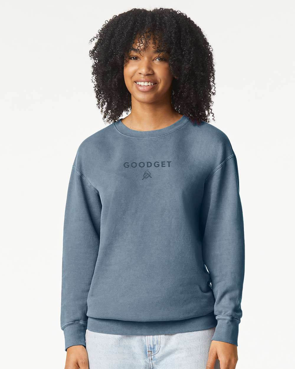 A washed blue crewneck sweatshirt by Good Get with a tone-on-tone logo, worn by a smiling woman.
