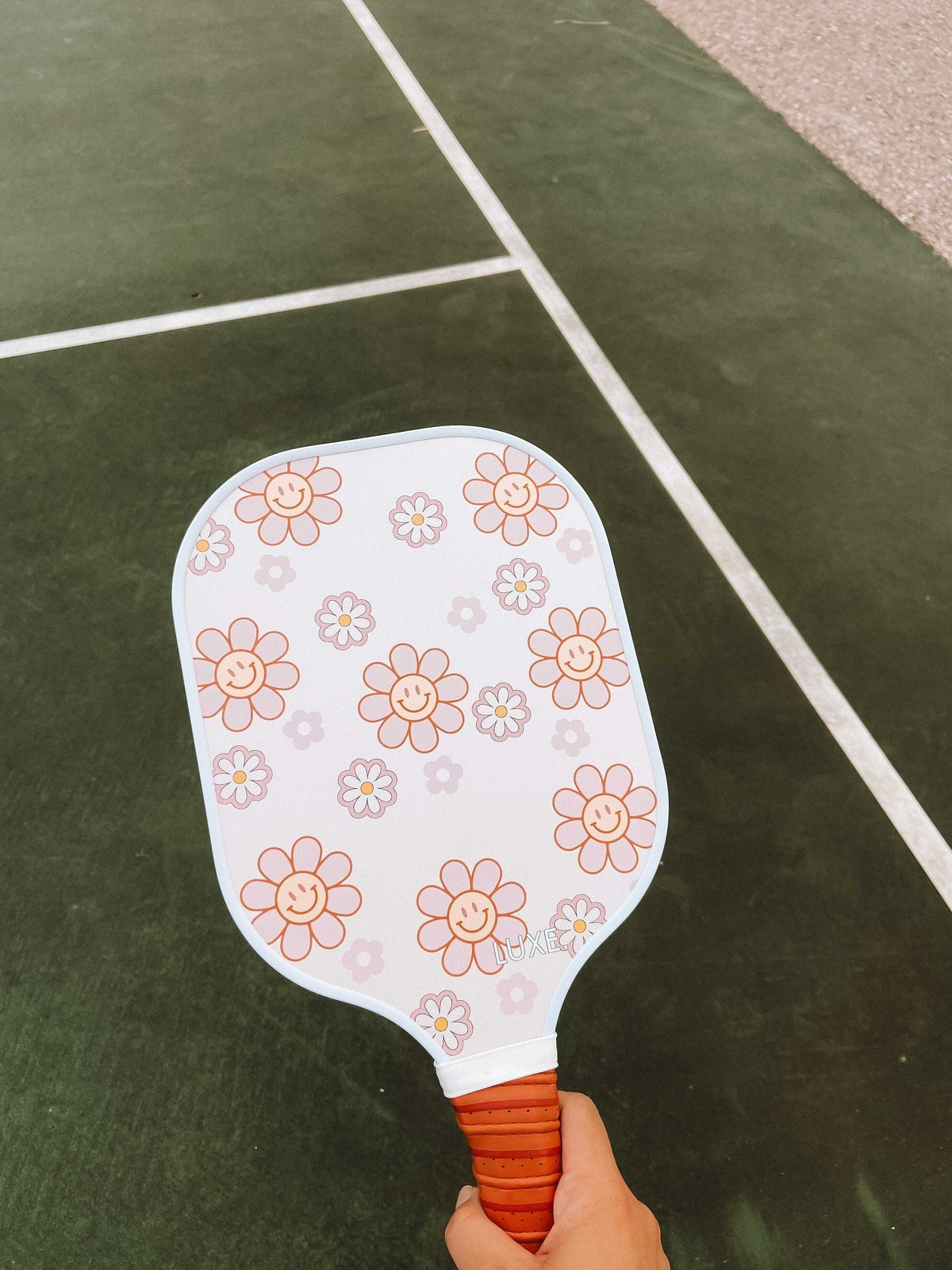 Person holding a white pickleball paddle with a pink flower design on an outdoor green court.