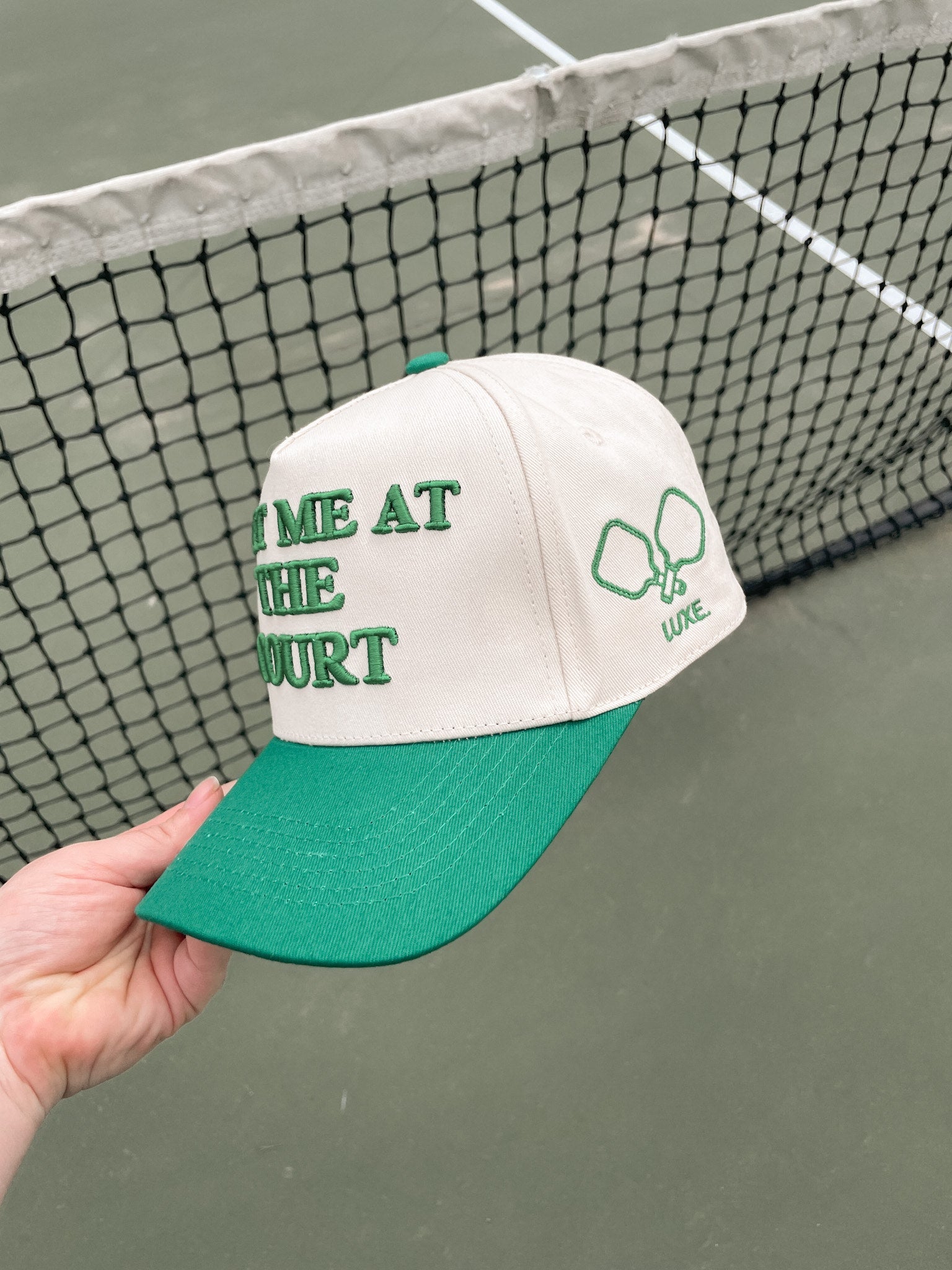 A beige and green baseball cap with "Meet Me at the Court" embroidered in green is held up against a blurred tennis court background. The cap has a green bill, and the logo of two crossed pickleball paddles is on the right side.