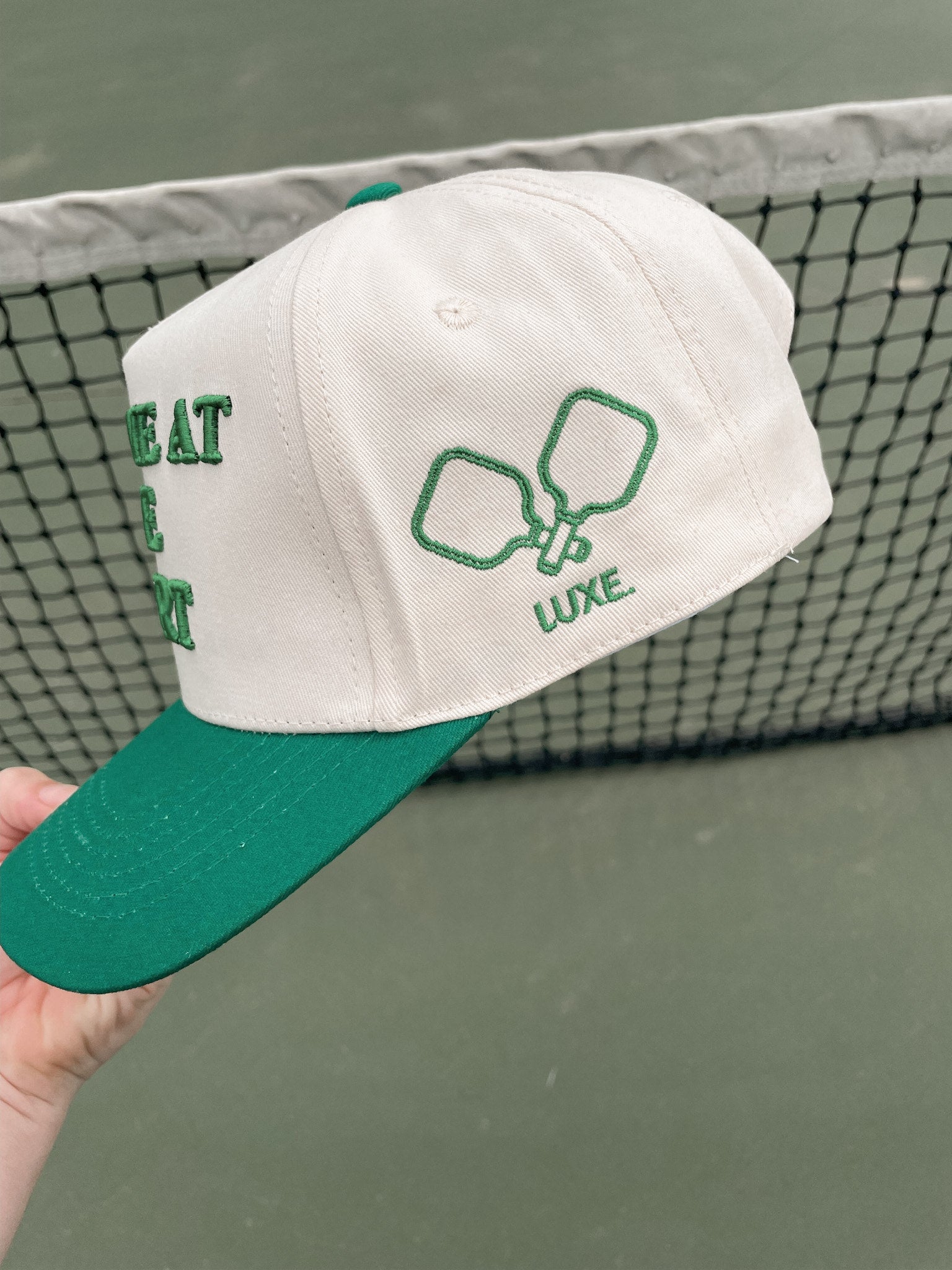 A white and green baseball cap with pickleball paddle embroidery is held up in front of a tennis net.