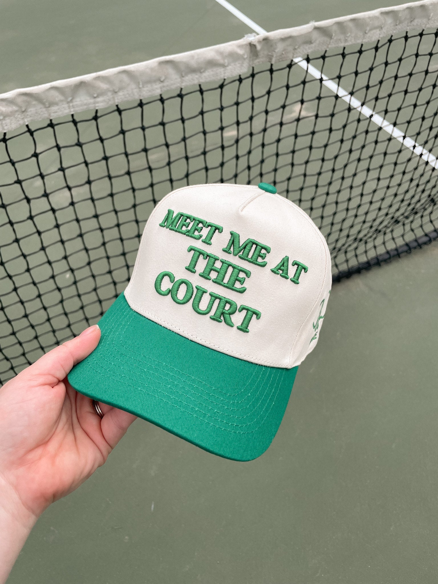 A hand holding a white and green baseball cap with "MEET ME AT THE COURT" embroidered in green, set against a tennis court backdrop.