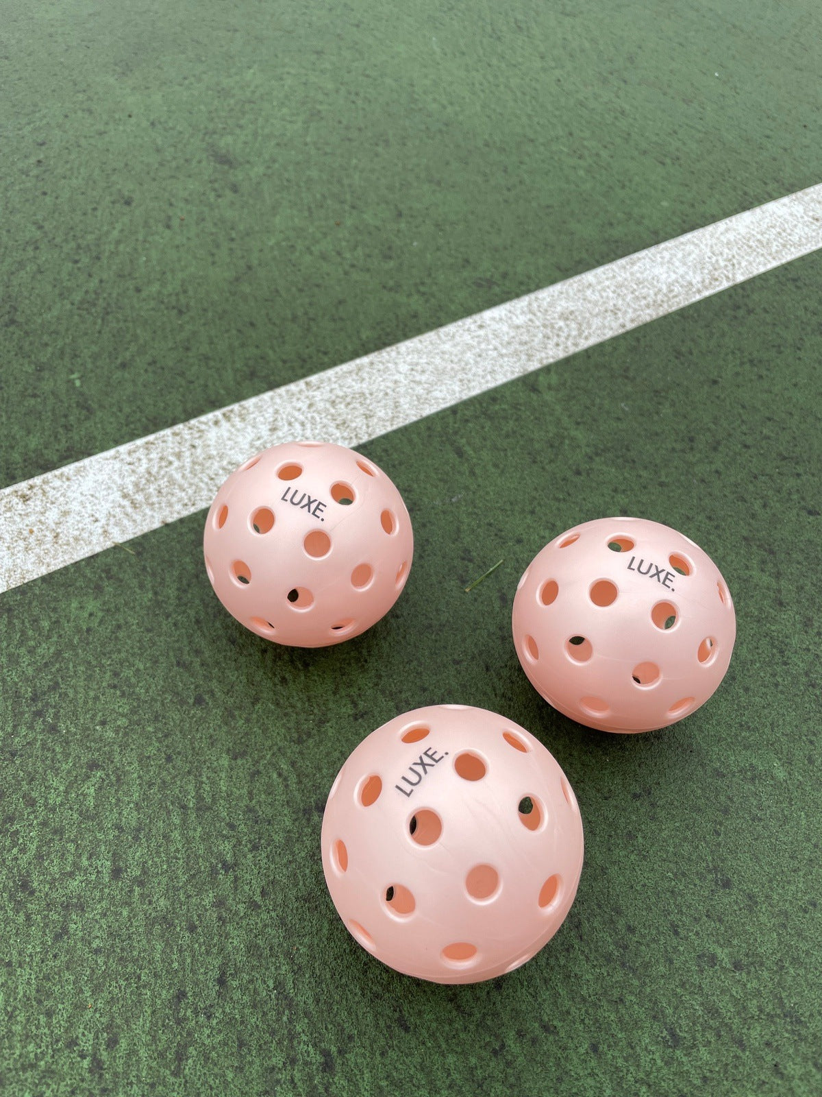 Three pink pickleballs with "LUXE" branding on a green and white court.